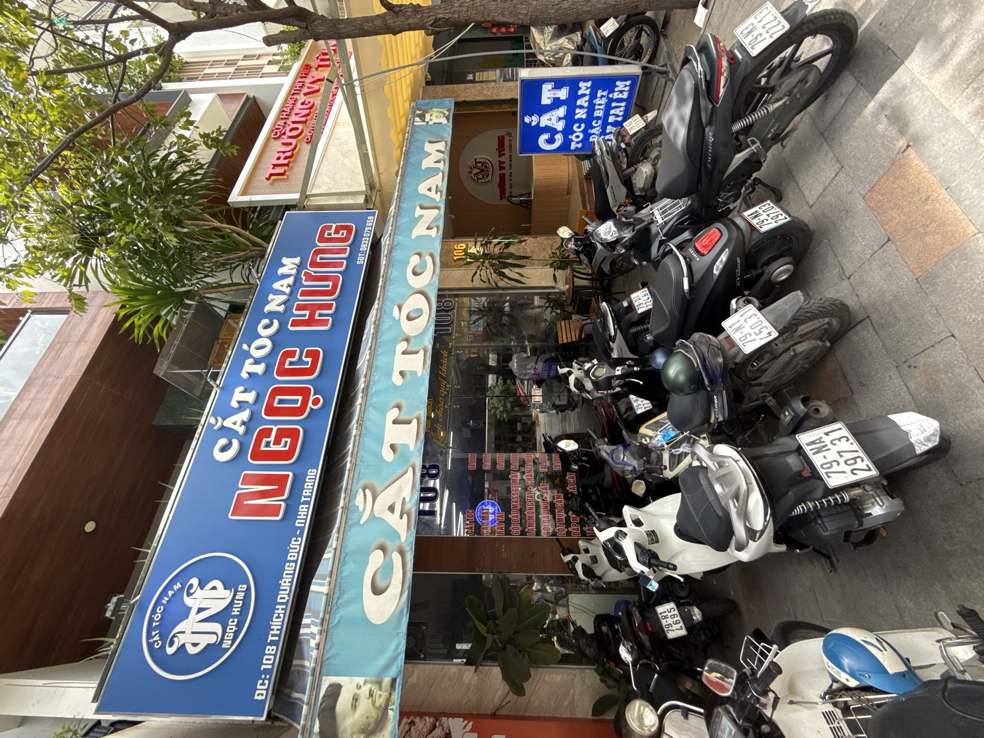 This is a street-level, eye-level shot of a barbershop called "CẮT TÓC NAM NGỌC HÙNG" located at 108 Thích Quảng Đức street in Nha Trang, Vietnam. The barbershop features a large blue sign with red lettering that reads "CẮT TÓC NAM NGỌC HÙNG". Below the main sign, there's a smaller awning with the text "CẮT TÓC NAM" in white lettering on a light blue background, and the number "108" is visible. A portrait of a man is displayed on the awning. The entrance to the shop is visible, with glass doors and a menu displayed inside. A variety of motorcycles are parked on the sidewalk in front of the establishment. Another shop, "TRƯỜNG VỸ TƯỜNG", is visible to the right. The street is paved, and trees and buildings line the street.