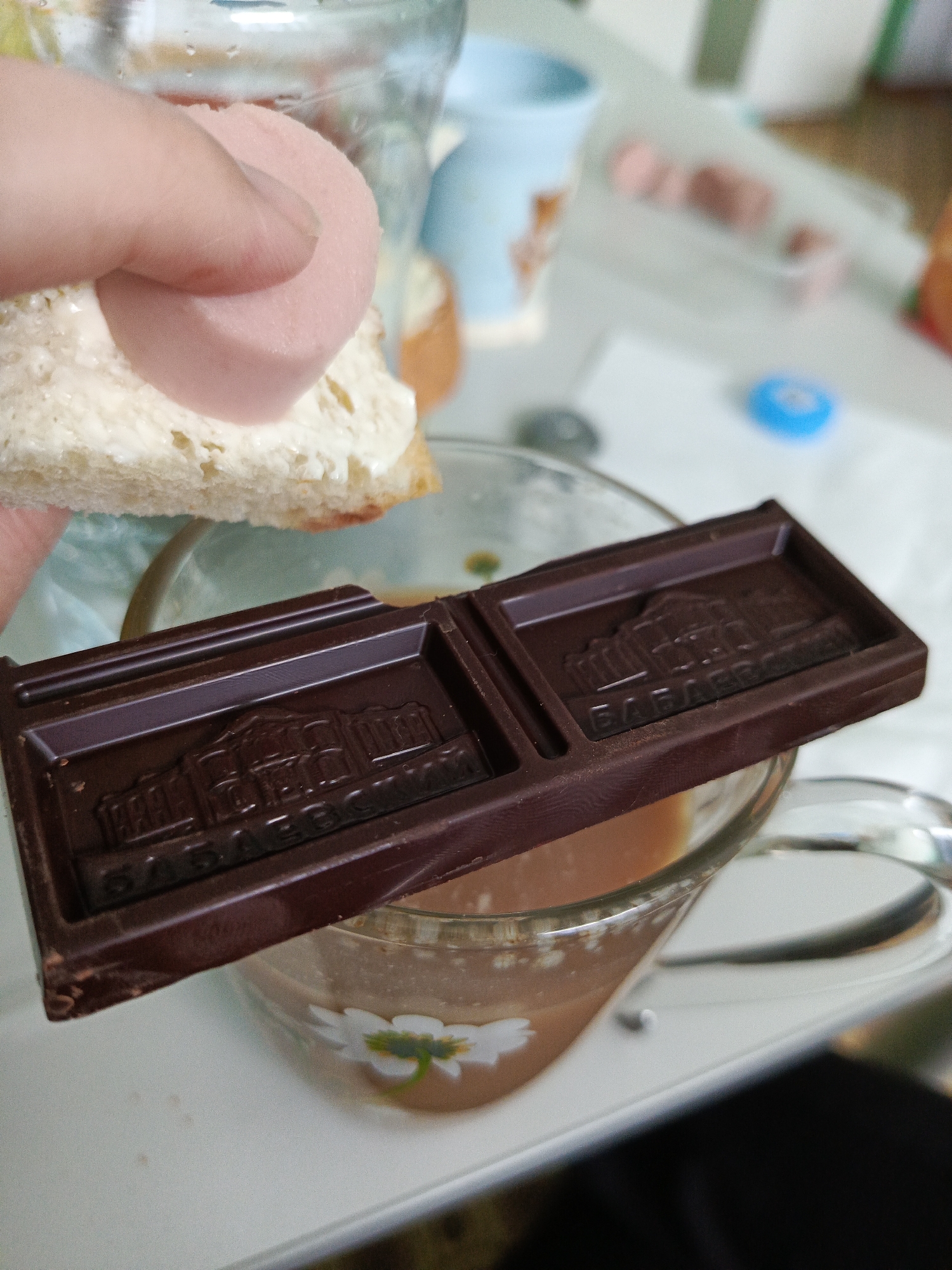 A human hand, with a thumb and forefinger, holds a piece of white bread spread with a creamy white substance, topped with a single, round slice of light pink processed meat. Below this, a dark brown chocolate bar, broken into two embossed segments, rests horizontally across the rim of a clear glass mug. Each chocolate segment features a raised architectural design and Cyrillic text, specifically "БАБАЕВСКИЙ". The glass mug contains a light brown liquid and has a white floral decal on its lower side. All items are positioned on a light-colored, flat surface, likely a table. The blurred background shows additional elements, including another clear glass container, a light blue mug, several similar pink processed meat slices scattered on the table, and a small blue circular object. The scene is located in Chaikovsky, Russia.
