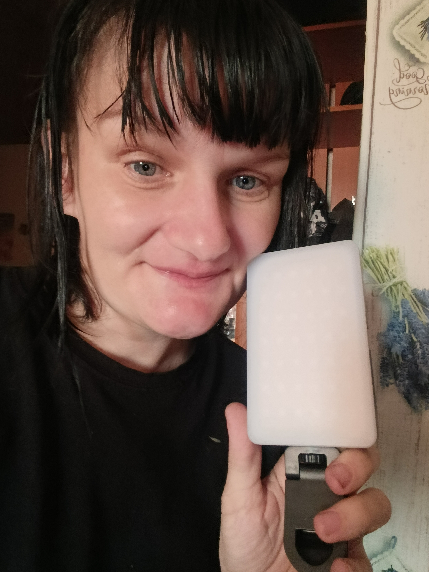 A woman is holding a white, rectangular object with a metal attachment in her right hand. She has short, dark hair with bangs and blue eyes. She is wearing a black t-shirt. The background includes a white wall with decorative writing and a small bouquet of blue and green flowers. The location is likely indoors, possibly in a kitchen or similar domestic setting. There is no direct indication of Фоки, Russia in the image.