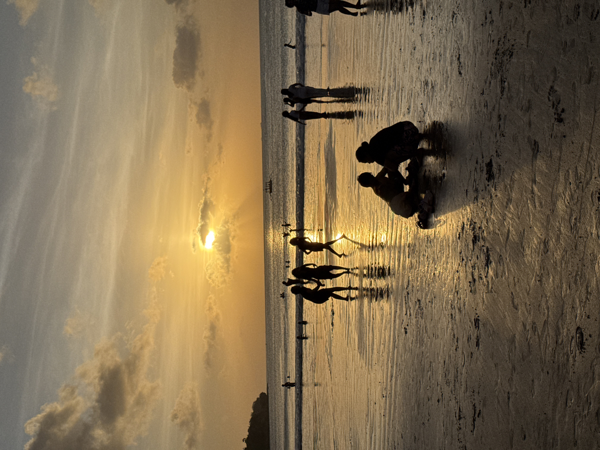 A dusk scene at Kuta Selatan, Indonesia, features several people silhouetted against a golden-hued sky and reflective beach. In the foreground, two individuals are crouched, possibly interacting with something on the sand. Farther back, groups of people are walking or standing, their forms elongated by the setting sun. The wet sand reflects the sunlight, adding to the atmospheric effect. In the distance, the horizon line is distinct, and small boats are visible on the water. Some clouds dot the sky. The sand shows footprints.