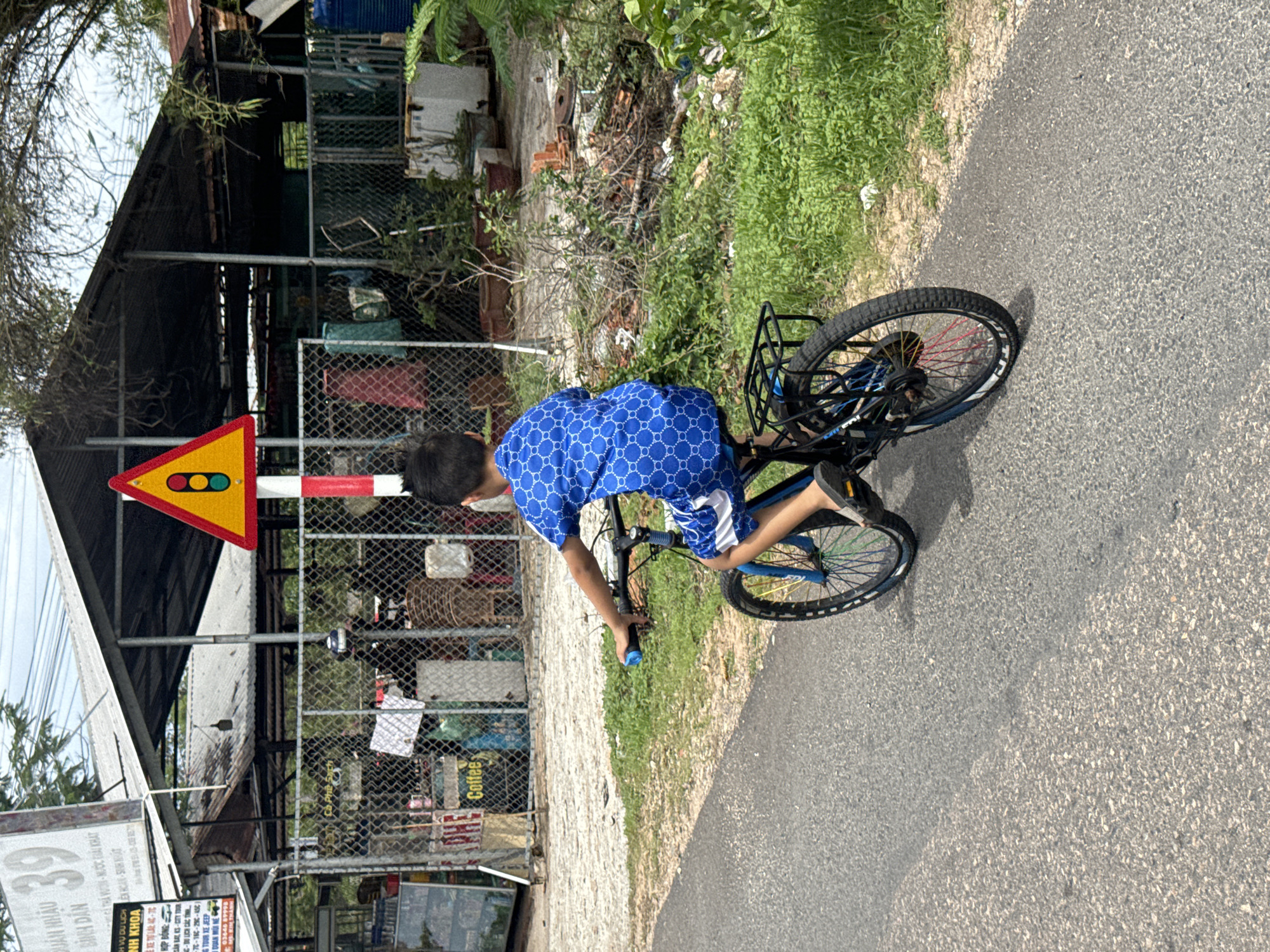 A daylight scene in Mũi Né, Vietnam, depicts a child riding a bicycle. The image is rotated 90 degrees counter-clockwise, showing a paved road on the right and a dirt path to the left. The child, viewed from behind, wears a blue shirt with a white circular pattern and shorts. They are riding a blue bicycle with a black rear rack and distinctively colored spokes in red, yellow, and green.
On the left side, a prominent yellow triangular warning sign, featuring a red, yellow, and green traffic light symbol, is mounted on a red and white striped pole. Behind the child and the sign are several simple structures, possibly shacks or workshops, partially hidden by chain-link fences and abundant vegetation including trees, bushes, and grass. The setting appears to be a semi-rural or industrial fringe area, bathed in clear, bright sunlight.
Visible text on various signs includes Vietnamese phrases like "QUẢN NHẬU", "BÌNH DÂN", "H VỤ DU LỊCH B", "NH KHOA", and the number "39". Other English text includes "SAN BAL KS-CITY TOUR", "CHC", numeric ranges such as "-70-160-250-450", "TOUR XE JEEP", and "998". A "Coffee" sign is also visible through a fence.