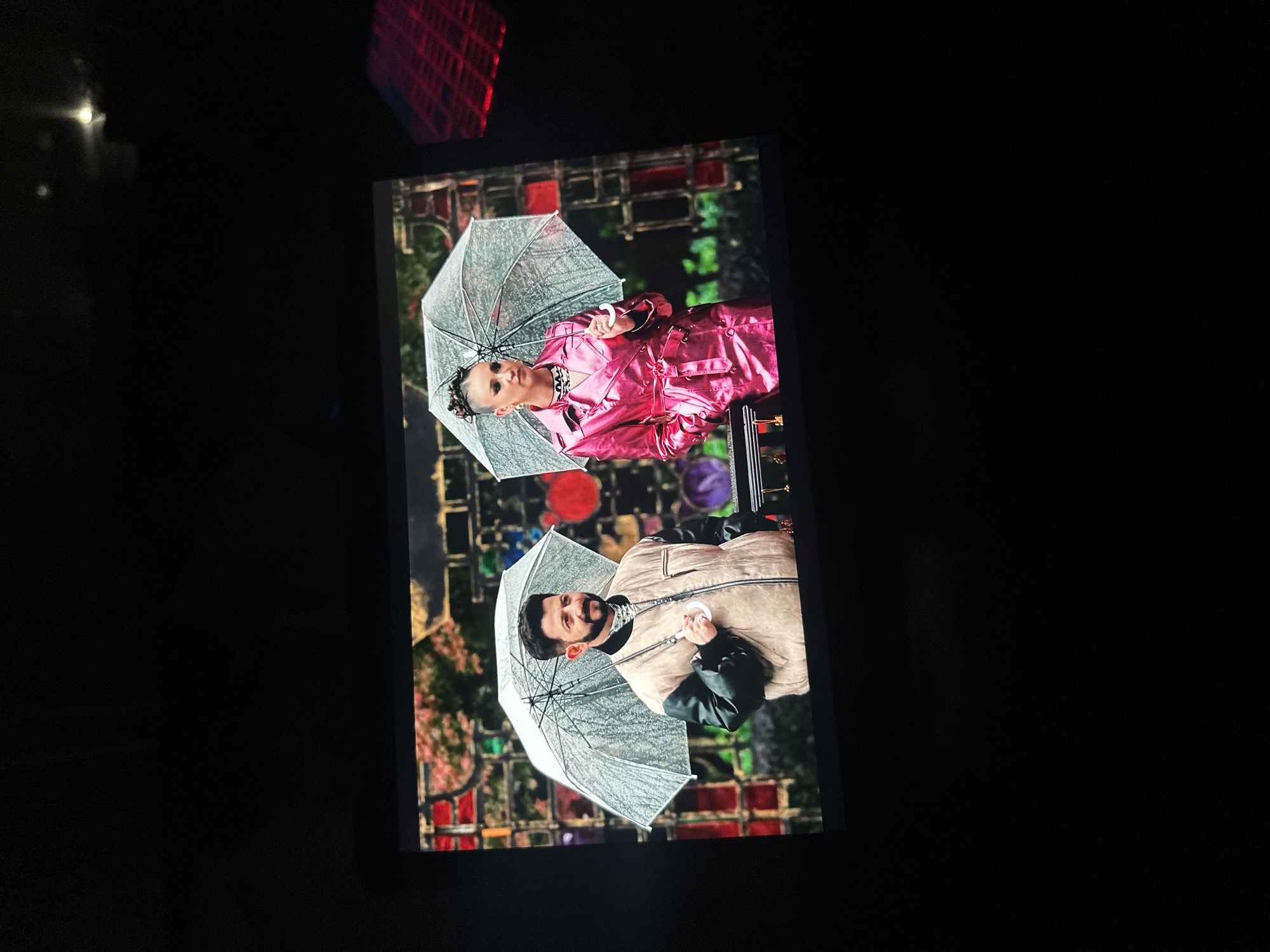 The image displays a screen showing two individuals holding clear umbrellas. On the left, a man with a beard wears a tan jacket with black sleeves and a black turtleneck. He is holding the umbrella with his right hand. On the right, a woman with an updo wears a bright pink trench coat. She is holding her umbrella with her right hand. They are standing in front of a background with what appears to be a mosaic or stained glass pattern. The screen is positioned in a dark environment. The location in the city of Novoaltaysk, Russia is not determinable from this image.