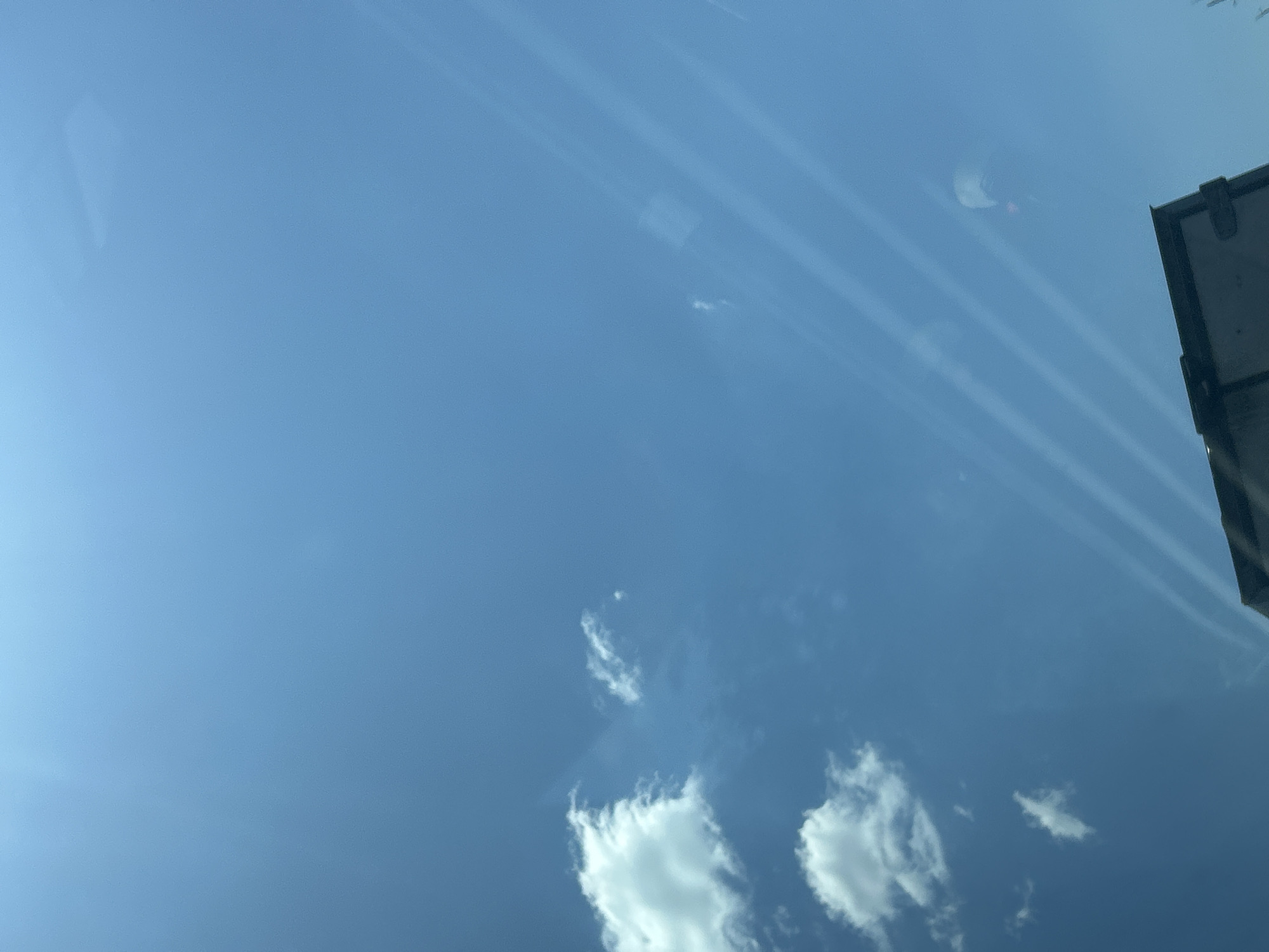 The image captures a bright, clear blue sky with scattered white clouds. The perspective suggests it was taken from inside a vehicle, looking upwards through a window, as evidenced by faint reflections and streaks that appear to be caused by the glass. In the lower right corner, the top edge of what appears to be a dark-colored vehicle or structure is visible, with a black rectangular shape that might be a cargo box or roof rack. The environment is a daytime sky, likely an outdoor setting, and the weather appears sunny and fair. There are no people or discernible actions taking place in the scene. The location is identified as Vakarel, Bulgaria.