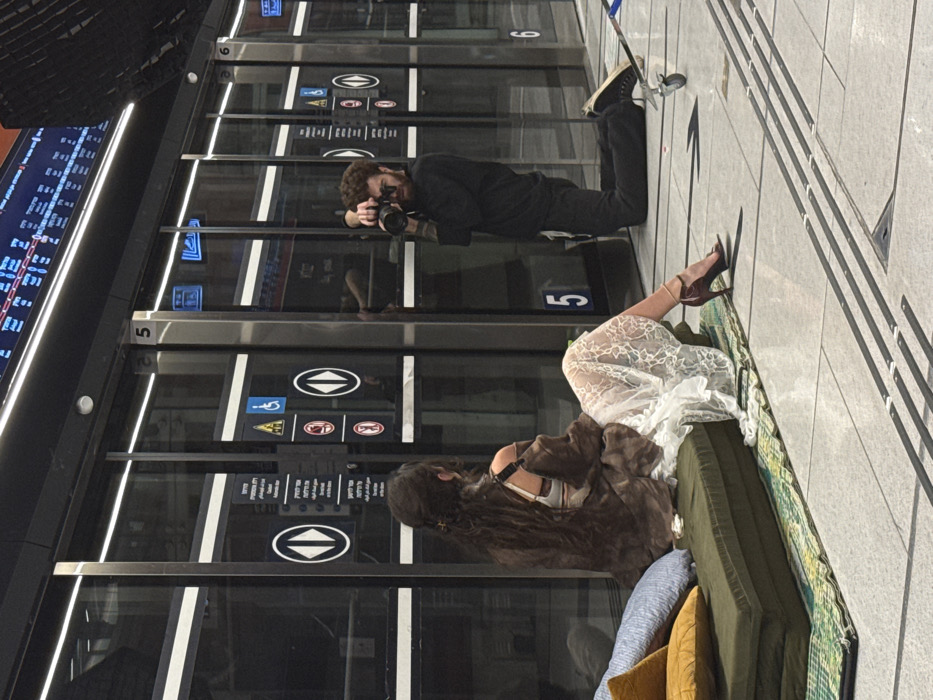 The image captures a photography session on a platform of the Tel Aviv Light Rail system in Tel Aviv, Israel.
In the foreground, a person with long, dark, wavy hair is seated facing away from the viewer, positioned on a stack of olive-green cushions placed on a green and yellow patterned mat. This individual is wearing a white lace skirt/dress, a white undergarment, and a loose brown shirt draped over their shoulders. A dark red high-heeled sandal with an ankle strap is visible on their extended left foot.
To the right, a male photographer with short, curly reddish-brown hair and a beard is kneeling on his right knee. He is clad in a dark long-sleeved shirt and dark trousers, holding a professional camera with a prominent lens, aimed directly at the seated person. Near his right knee, a small, silver-colored wheeled piece of equipment rests on the tiled floor.
The background features modern platform screen doors constructed of glass and brushed metal frames. These doors display various symbols and text, including circular white arrows on black backgrounds, a blue wheelchair accessibility icon, red prohibition symbols, and signage with Hebrew text. Above the doors, a long, horizontal digital display panel shows dynamic information, also predominantly in Hebrew. The platform floor is light-colored tile with embedded dark gray lines and a black directional arrow.