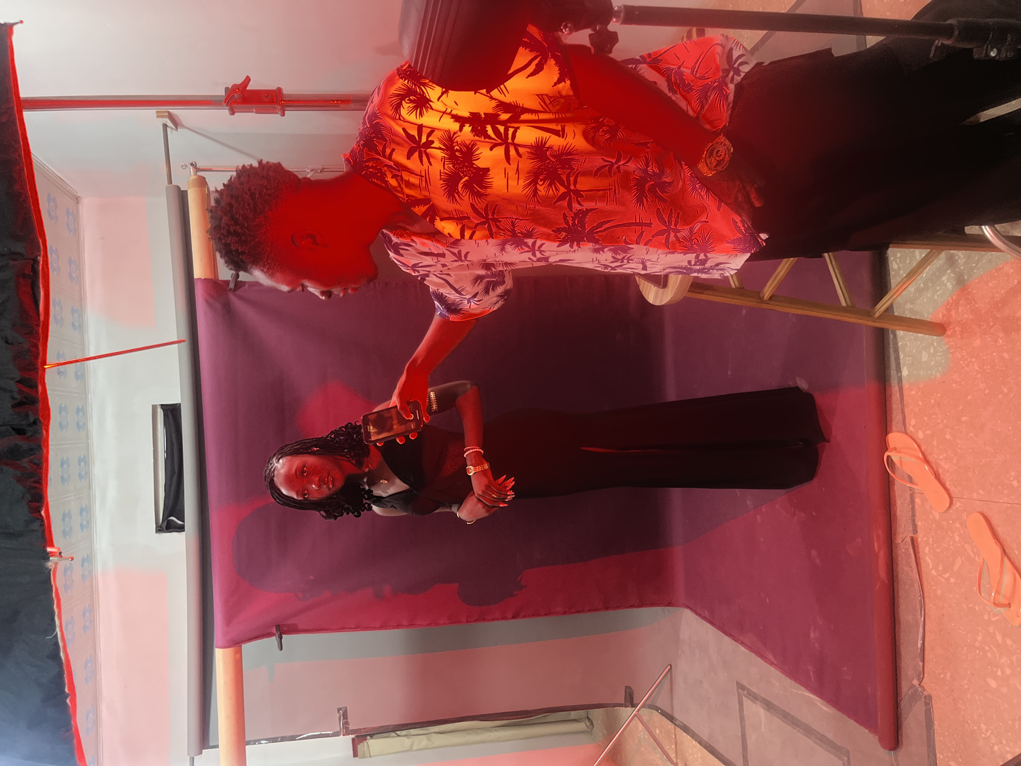 In a brightly lit room with red lighting, a young Black woman stands facing left, wearing a black dress and holding a cell phone in her right hand, looking into it. She has braided hair and is wearing bracelets and a necklace. To her right, a young Black man stands facing her, holding a cell phone in his right hand, appearing to be taking a picture or recording. He has short, curly hair and is wearing a floral shirt and dark pants. A wooden stool is positioned between them. In the foreground, a pair of orange flip-flops are visible on the floor. The background consists of a grey backdrop with a burgundy fabric draped over a stand. The overall setting suggests a photo studio or a similar space for creative work, likely within Gura Top, Nigeria, indicated by the request's context.