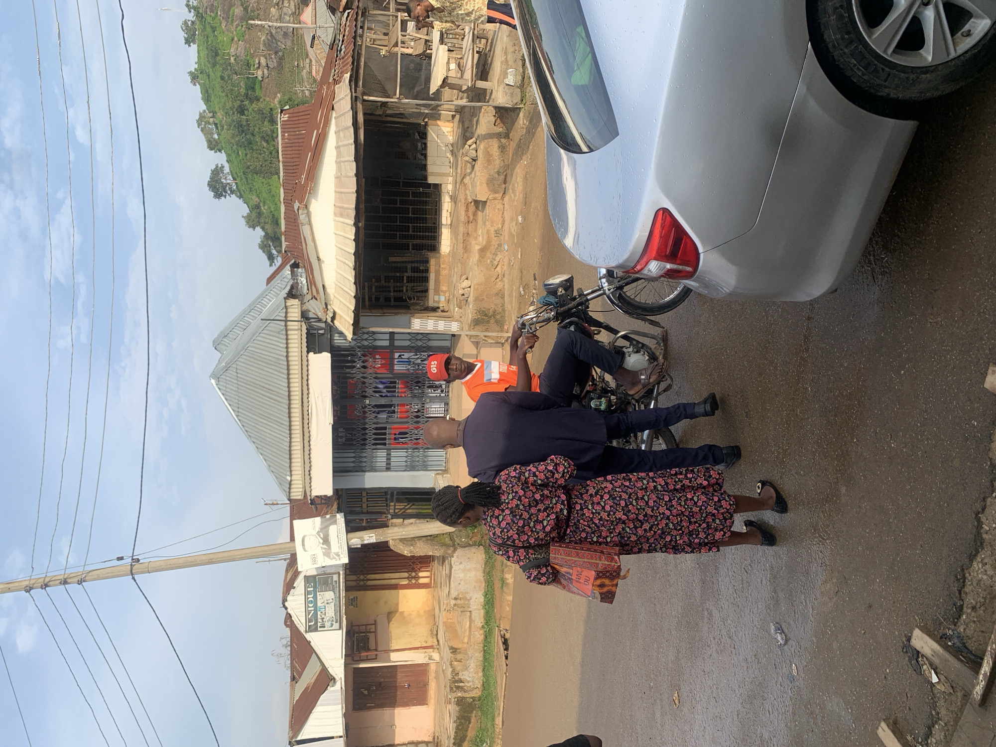 The image depicts a street scene in Abuja, Nigeria, with several individuals and vehicles. In the foreground, a silver car is parked on the wet, asphalt road. To the left of the car, a man in a dark suit stands with his back to the camera, facing a woman in a floral print dress who is carrying a patterned tote bag.
In the middle ground, a motorcycle is positioned on the road. A man wearing an orange shirt and a red cap with "G15" printed on it is seated on the motorcycle, appearing to be engaged in a conversation or transaction with the man in the suit. Behind them, several storefronts with corrugated metal roofs line the street. One of the storefronts has a sign that reads "UNIQUE." The buildings appear to be constructed with concrete and metal.
In the background, a lush green hillside with scattered trees and rocks is visible under a cloudy sky. Power lines are strung across utility poles, typical of urban infrastructure. The overall environment suggests a somewhat overcast day, with evidence of recent rain on the road.