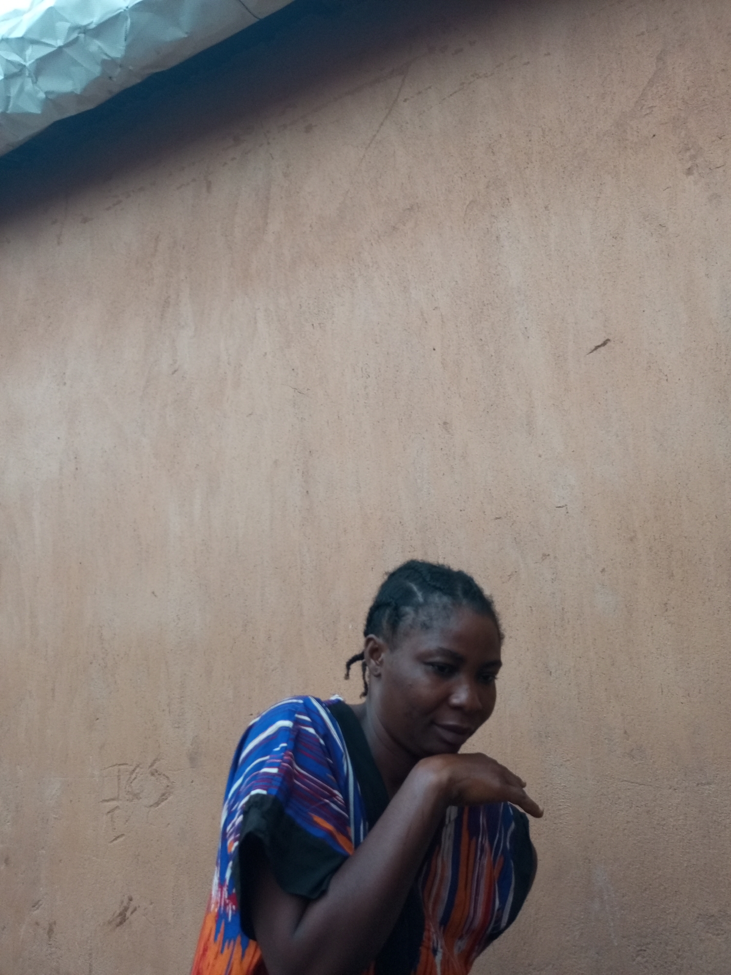 A woman with dark hair braided in cornrows is sitting against a tan, textured wall with some faint markings. She is wearing a vibrantly colored top with horizontal stripes in blue, white, orange, and black. Her right arm is bent, with her hand resting on her chin, and she is looking to her right. In the upper left corner of the image, a crumpled metallic material is visible, possibly part of a roof or awning. The overall lighting suggests it is daytime. The scene appears to be outdoors, likely in a residential or utilitarian area in Tempure, Nigeria.