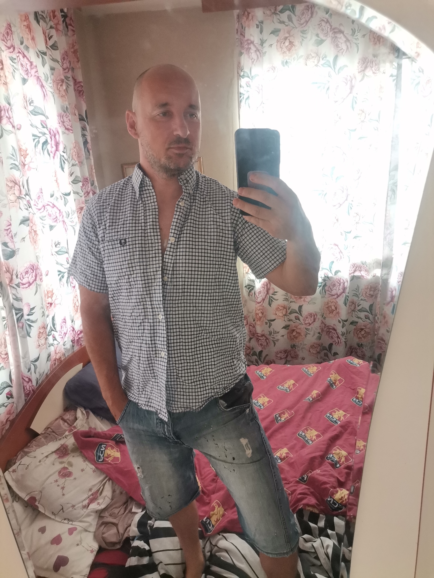 A fair-skinned, bald man with a stubble beard stands indoors, reflected in a mirror. He wears a short-sleeved, button-down plaid shirt and denim shorts with his right hand in the pocket. He holds a black mobile phone in his left hand, presumably taking the picture. Behind the man, a bed with patterned bedding is visible. The room is decorated with floral curtains, and the location is Unknown, Unknown.