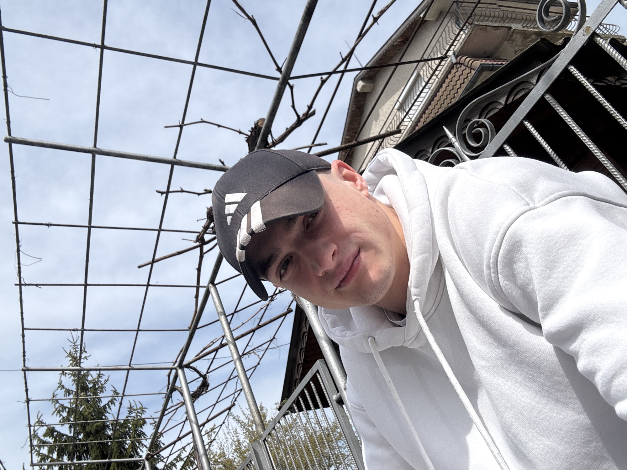 A young man wearing a white hoodie and a black baseball cap with white stripes on the brim is looking directly at the camera. He is positioned beneath a metal trellis structure with bare vines. Behind the trellis, a green evergreen tree can be seen. To the right, part of a building is visible, with ornate metal railings and a tiled roof. The sky is overcast. This image appears to be taken in Topoli, Bulgaria.