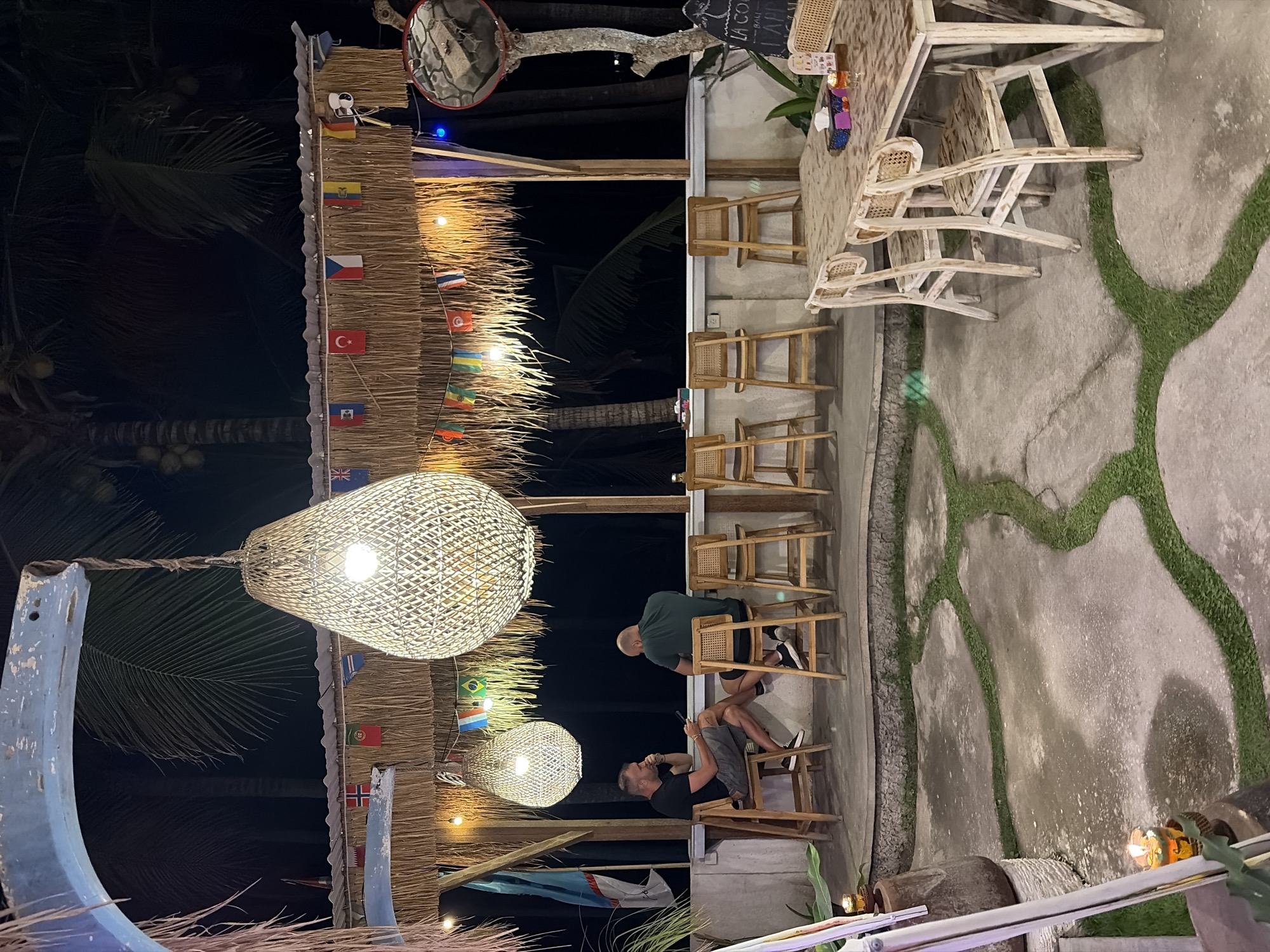 The image depicts an outdoor bar area at night, likely in Nusa Penida, Indonesia. Two men are seated at a bar, one facing left and the other facing right, appearing to be in conversation. The bar is constructed of natural materials like thatch and wood, with bamboo chairs and stools. Several flags of different nations are strung across the thatched roof, suggesting an international clientele. Large, woven lamps hang overhead, providing warm lighting. In the foreground, a concrete patio is interspersed with strips of grass, creating an organic pathway. A wooden table with chairs is set up to the right, with a candle and tissues on it. The background is dark, with the silhouettes of palm trees visible, indicating a tropical location. The overall ambiance is relaxed and casual.