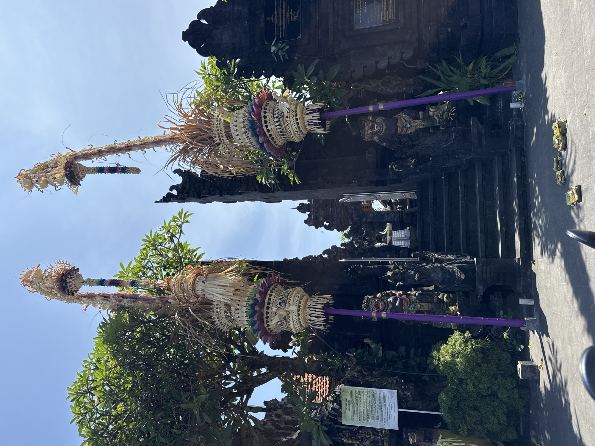 Two penjors, traditional Balinese bamboo poles decorated with woven coconut leaves and offerings, stand in front of a dark stone gate in Nusa Dua, Indonesia. The gate is adorned with carvings of deities. Trees and shrubs surround the gate and penjors. A sign is visible to the right of the frame. The sky is partially visible at the top of the image.