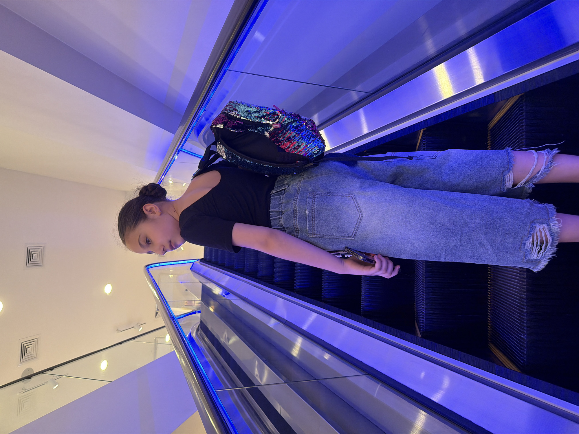 A young girl with dark hair pulled back in a bun, wearing a black t-shirt and ripped denim capris, rides an escalator. She carries a sequined backpack and a cell phone. The escalator, illuminated by blue LED lights, appears to be in a modern indoor public space, possibly a shopping mall. The background shows ceiling details and ventilation grates. The environment is well-lit and clean. There is no visual information to suggest this location is in Yerevan, Armenia.