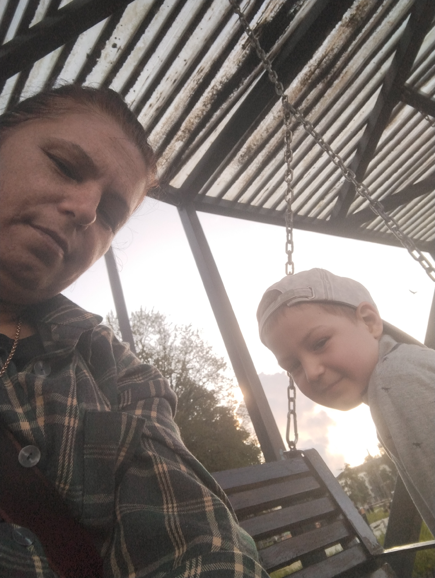 In Kingisepp, Russia, a close-up selfie is taken from a low angle, capturing two individuals on a swing set. The person in the foreground, to the left, is an adult with light brown hair pulled back, wearing a dark green and tan plaid shirt over a dark garment and a gold chain necklace. Their face is partially obscured, but they appear to be looking down with a neutral expression. To the right, a young boy wearing a grey baseball cap and a grey shirt with a subtle dark pattern is closer to the camera. He is looking towards the camera with a slight smile. Both individuals are on a wooden swing seat, suspended by metal chains from a structure with a corrugated metal roof overhead. The background shows trees and a bright, overcast sky, suggesting an outdoor setting, likely a park or playground, in Kingisepp.