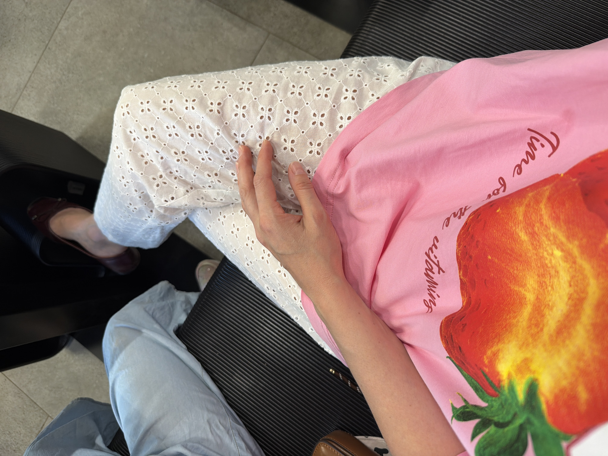 A high-angle shot captures a person seated on a black chair with striped upholstery, with their legs crossed. The person's attire includes white eyelet-patterned pants, and a pink top featuring a large image of a strawberry and partial lettering that reads "Time for the...". Visible on the left foot is a dark brown flat shoe. A light blue garment is visible below the seat. The floor is covered with gray tiles. It is not possible to determine the location as Izhevsk, Russia from this image.