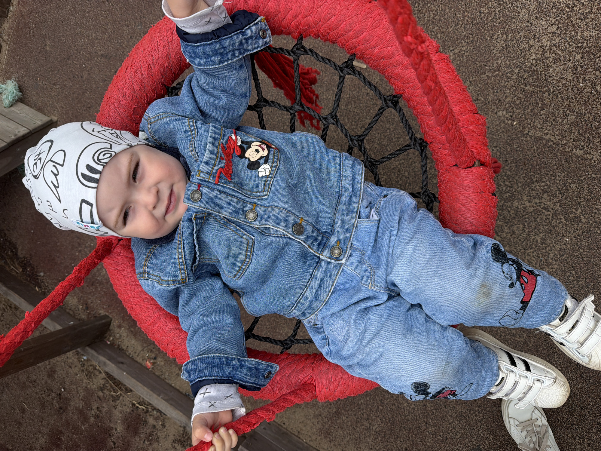 A toddler is sitting on a red, netted swing. The child is wearing a denim jacket and jeans with Mickey Mouse designs, a white beanie with black doodles, and white sneakers. The swing is suspended by thick red ropes, and the ground beneath appears to be a rubberized surface typical of a playground. Wooden planks are visible in the background. This scene is located at a playground in Votkinsk, Russia.