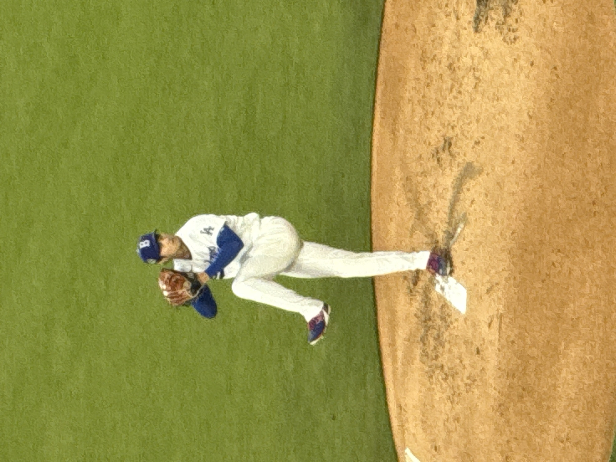 The image shows a baseball player in a white uniform with blue accents, including a Los Angeles Dodgers cap. He is in the middle of a throwing motion, with his glove up and focused. He is positioned on the pitcher's mound, which is made of dirt. The surrounding field is green. The image may be taken at a stadium in Los Angeles, United States of America.
