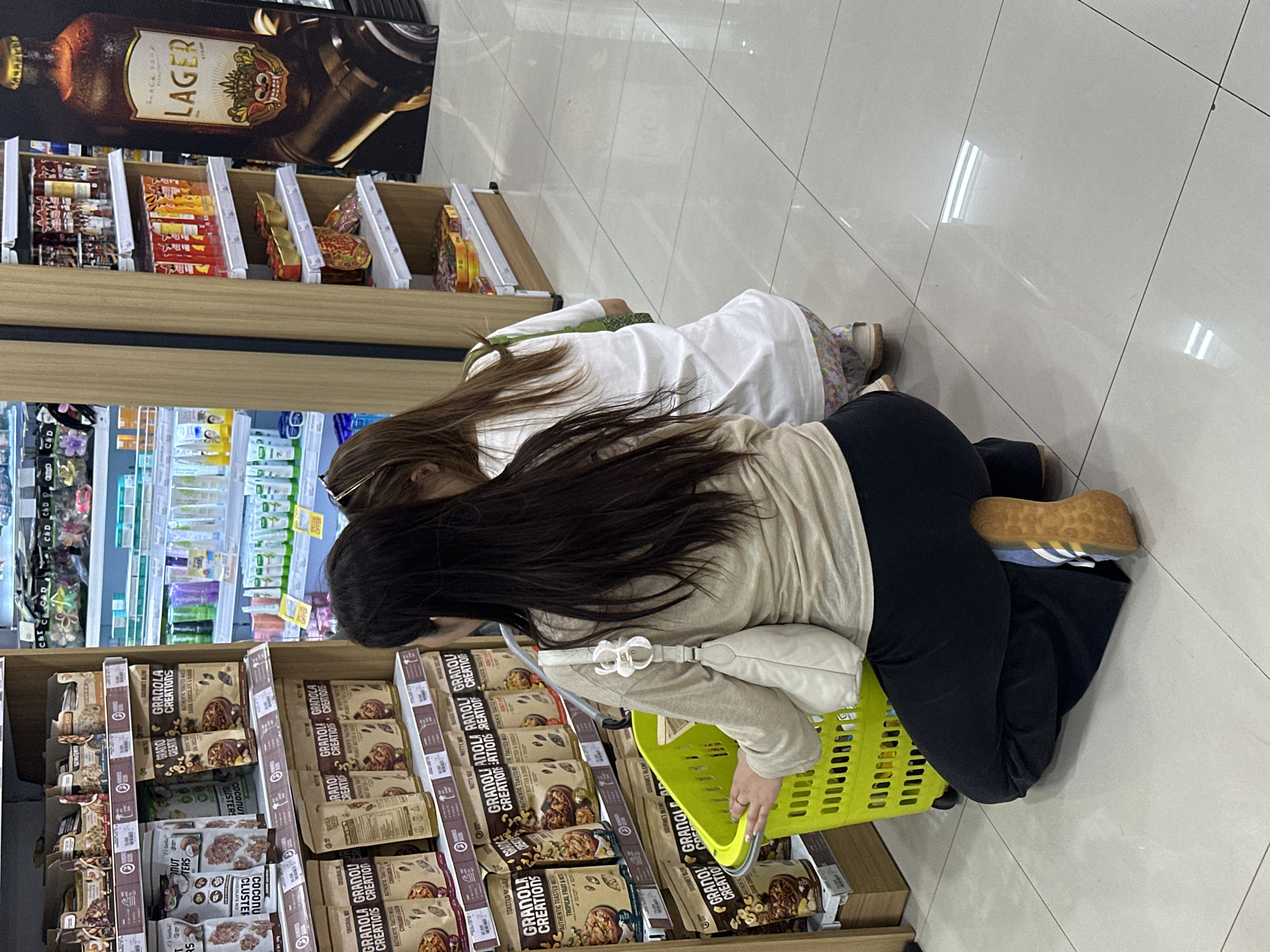 Two individuals, likely a mother and child, are seen in a grocery store aisle in North Kuta, Indonesia, examining products on a shelf. The primary subject, an adult woman, is crouched down, her hands resting on a bright yellow shopping basket. She is wearing a beige long-sleeved shirt and black pants, with a white handbag slung over her shoulder. Beside her, a younger person, presumably a child, is also crouched and looking at the shelves. They are wearing a white t-shirt and floral patterned leggings. The shelves are stocked with various packaged food items, including granola, and other assorted products. The environment appears to be a well-lit retail space with tiled flooring.