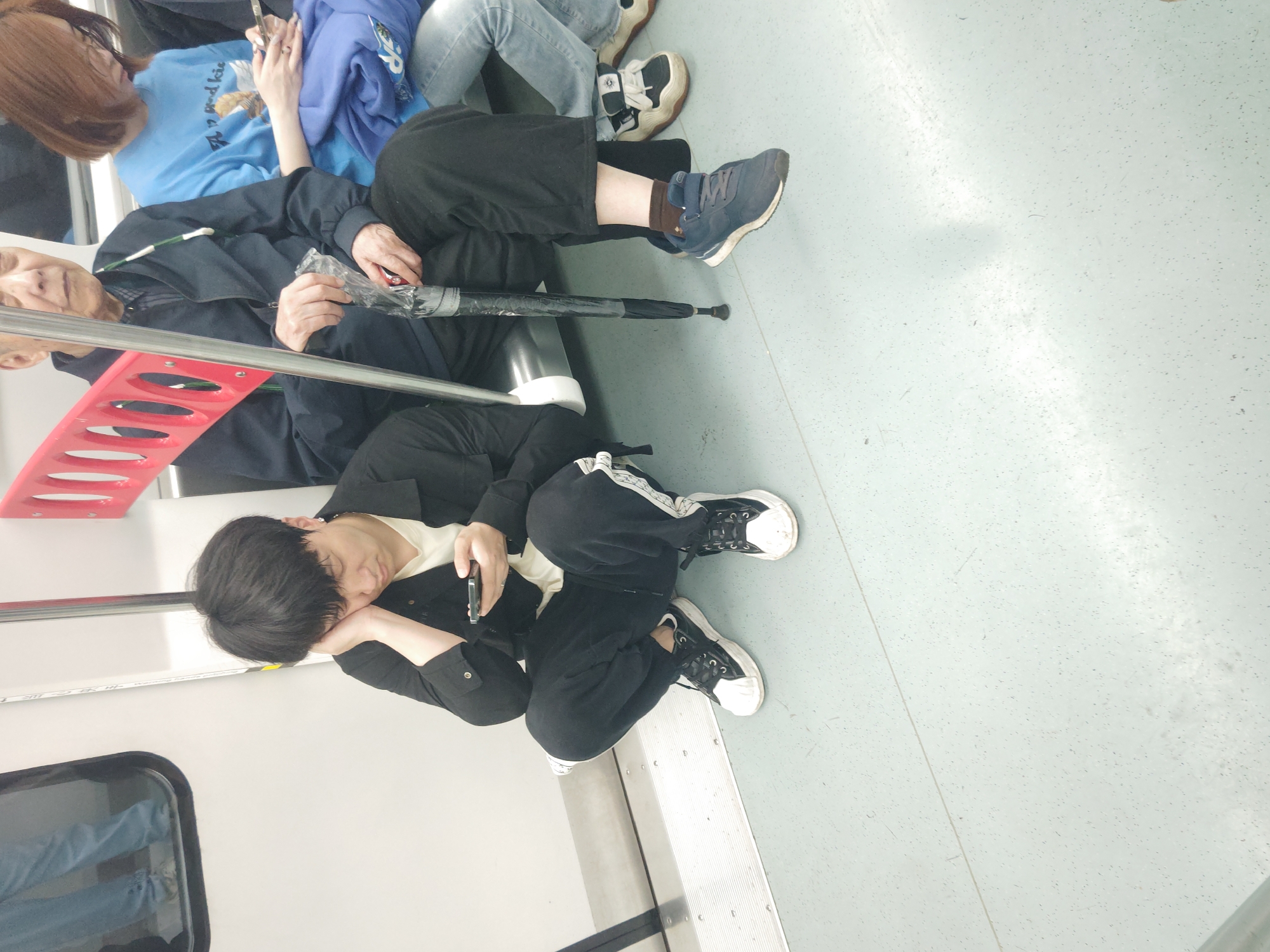 The image shows the interior of a subway car in a city of Unknown, Unknown. There are at least four people visible in the scene. An elderly woman sits on the left, wearing a dark blue jacket. Next to her is a younger person wearing a light blue hoodie, blue jeans, and white shoes. In front of her, a person is crouched down using the subway wall for support. The person is wearing a black jacket, cream undershirt, black pants, and white and black sneakers. They are holding a phone and resting their head on their hand as if they are sleeping or tired. An umbrella rests in between two seats. The floor is light gray with speckled marks. The subway car has red handrails and the interior is lit by overhead lights. There is a window on the left with a view of another building outside.