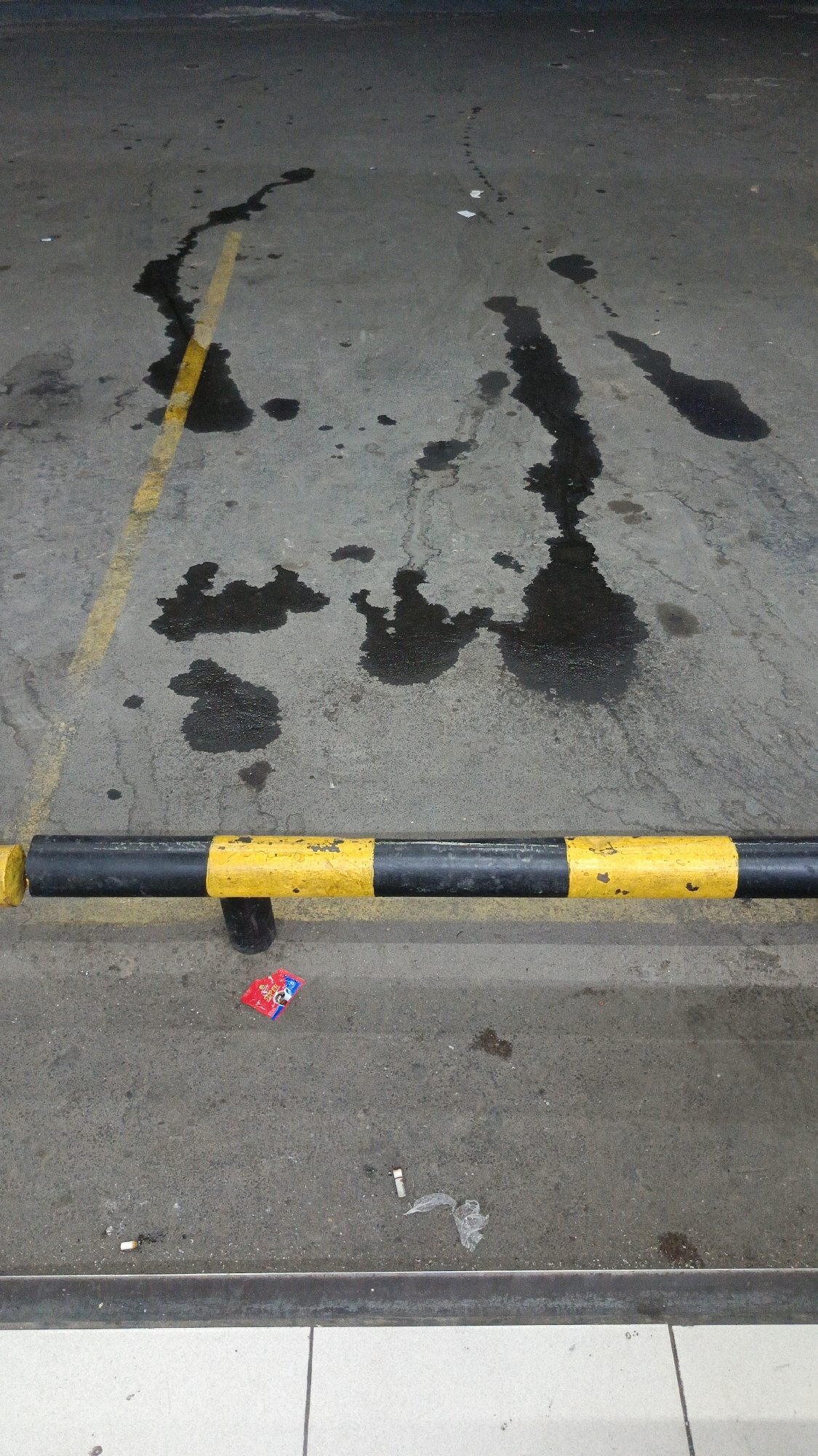 The image shows a section of asphalt pavement, possibly in a parking lot, at street level. A yellow and black striped barrier runs horizontally across the lower portion of the frame, indicating a curb or edge. The pavement is wet in several areas, with dark, irregular patches of liquid, likely oil or water. A yellow line, possibly a parking space marker, runs diagonally from the upper left towards the center. On the ground in front of the barrier, there is a small, red and blue wrapper, possibly a candy or gum package, and a few cigarette butts. The overall environment appears to be urban, with the pavement showing signs of wear and tear. The location is specified as Margajaya, Indonesia.