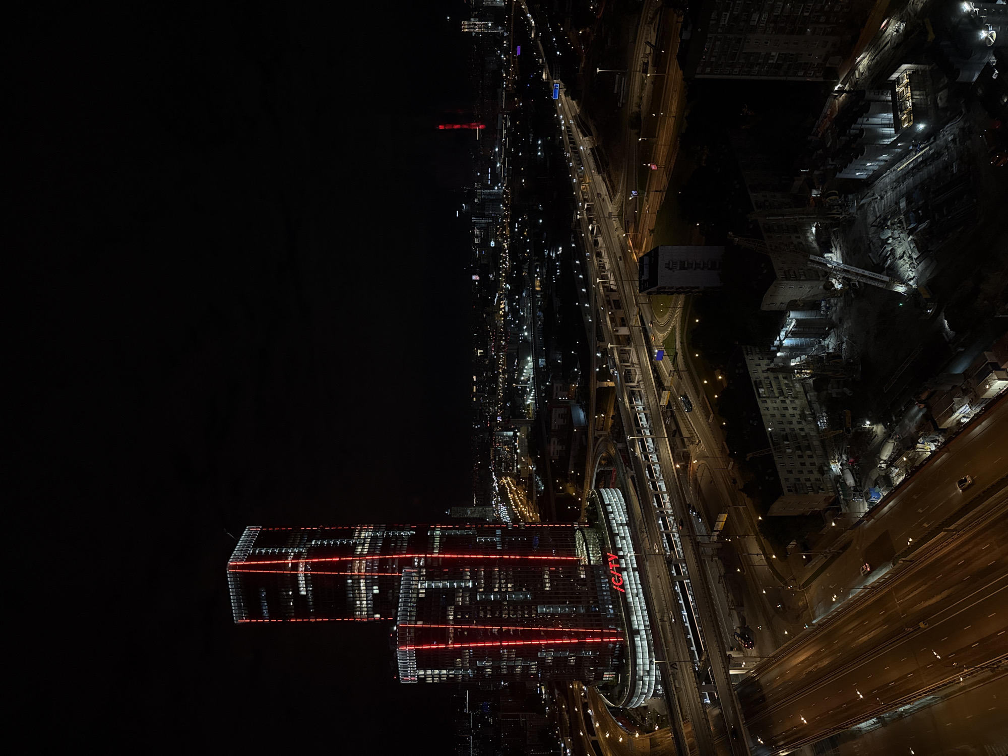 The image presents a high-angle, vertically oriented view of an urban landscape at night. The scene is illuminated by countless artificial lights against a dark sky.
In the lower-left portion, two striking modern skyscrapers dominate, featuring numerous illuminated windows and distinctive horizontal red LED strips that wrap around their facades. The prominent red text "ICITY" is visible on one of these buildings, serving as a key location identifier.
To the right of these tall structures, an extensive network of multi-lane highways and elevated roads stretches across the city. These roads are brightly lit by streetlights, with visible streaks of light indicating moving vehicle traffic. Parallel to some of the highways, an illuminated railway line or elevated transit system is also apparent.
Further to the right and extending into the upper parts of the image, various other urban buildings and infrastructure are visible. A significant portion reveals a large construction site, brightly illuminated by floodlights, showcasing machinery and excavated areas.
The overall impression is that of a bustling, active city thriving at night. The weather appears clear. The extensive artificial lighting highlights the scale and intricate network of the city's infrastructure. The scene depicts a typical night in a large urban center, with ongoing activity. The location is Khimki, Russia.