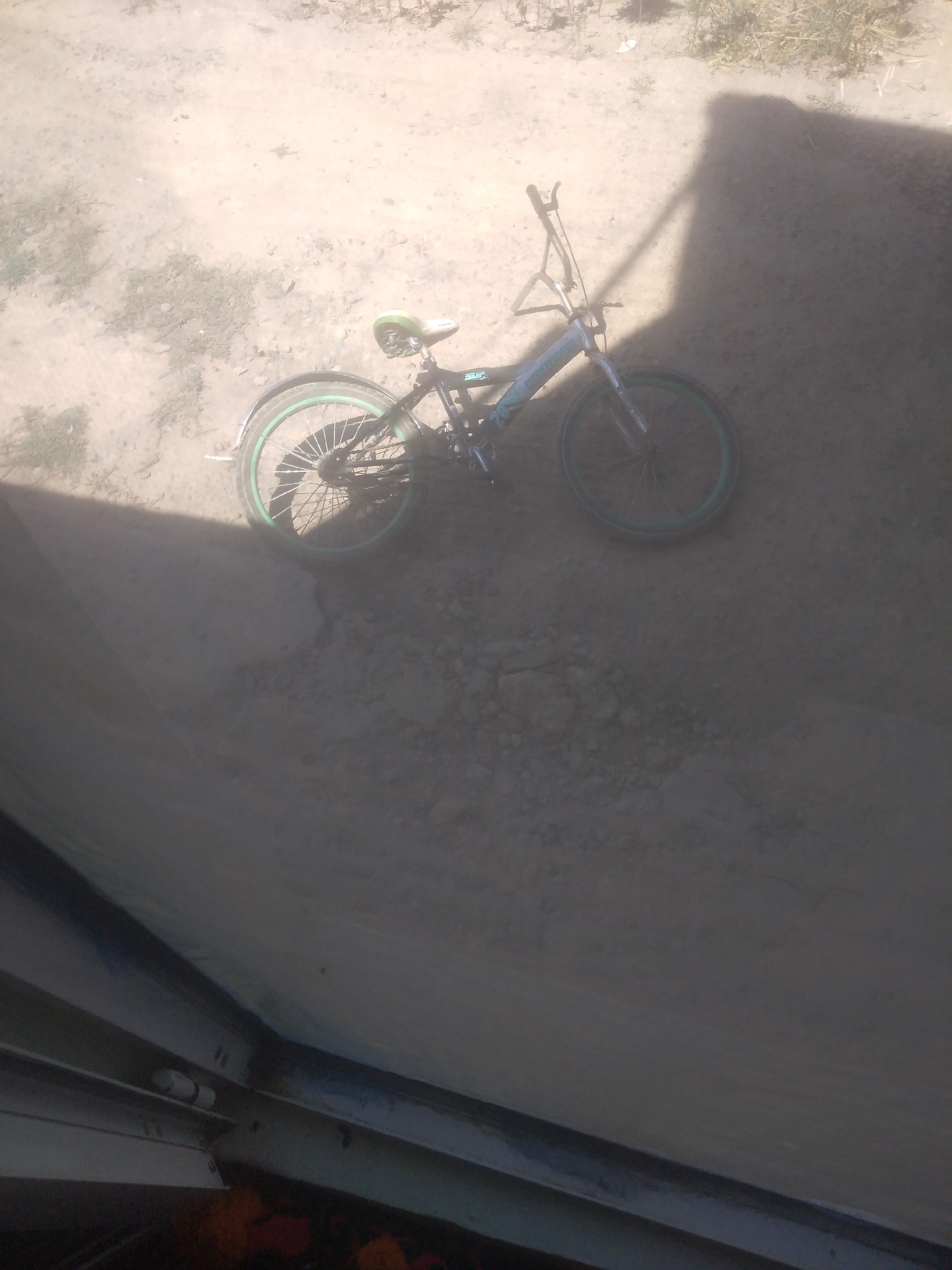 An outdoor, low-angle shot captures a bicycle leaning against an unseen object, casting a long shadow across a dirt ground. The bicycle is dark-colored with green accents on the rims and seat. The front of the bicycle faces towards the right of the frame, and its handlebars are visible. The background shows dry dirt with sparse vegetation. In the foreground, a dark, reflective surface partially obstructs the view, with hints of orange and red beneath it. The scene is under bright sunlight, suggested by the harsh shadow.