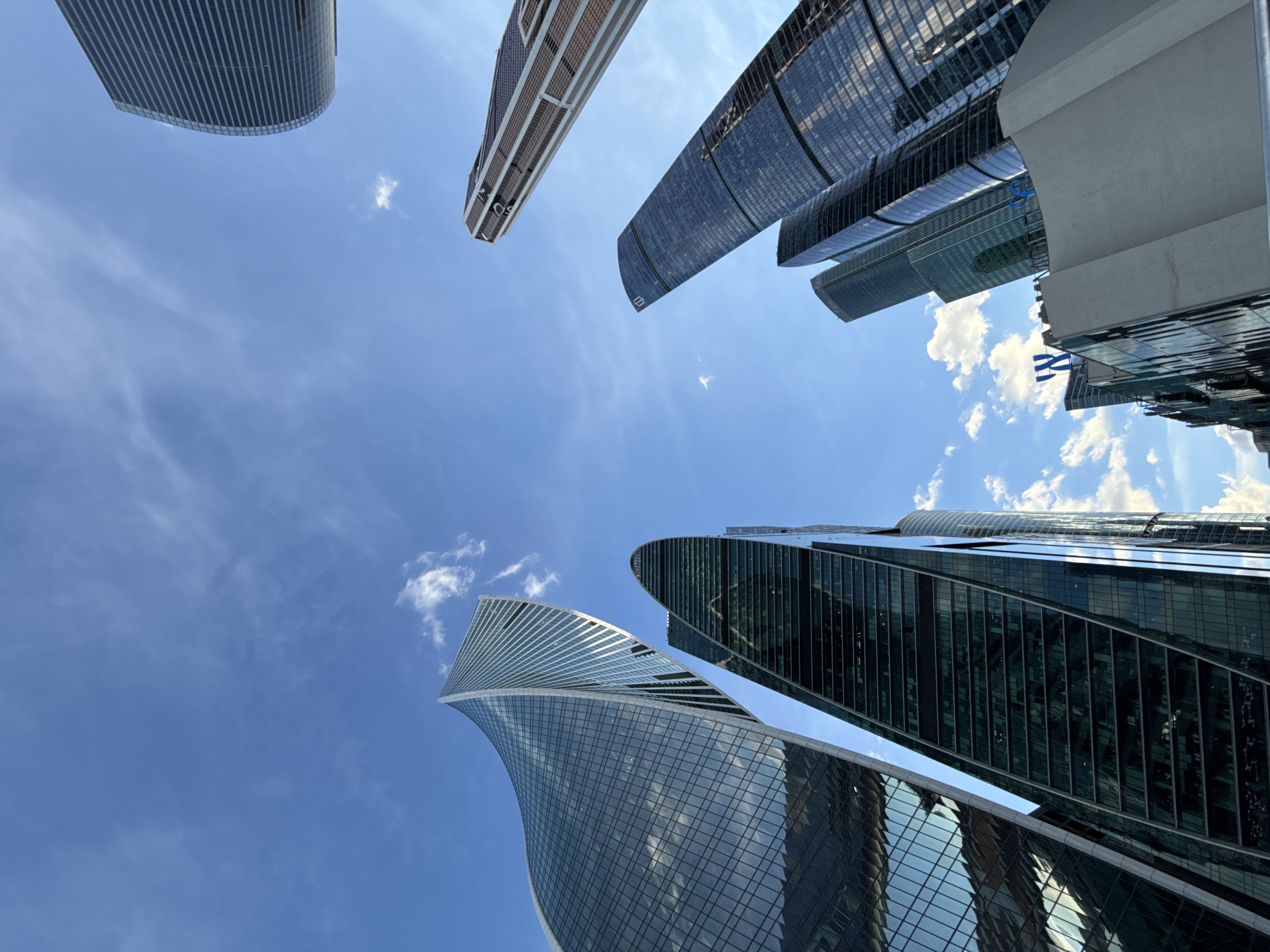 This is a low-angle shot of several modern skyscrapers against a clear blue sky with scattered white clouds. The buildings are tall and appear to be made primarily of glass and steel, with geometric and curved architectural designs. The perspective emphasizes the height and scale of the structures, making them seem to reach towards the sky. The sun is bright, suggesting it is daytime, likely late morning or afternoon. The scene is an urban environment, possibly a business district or a city center known for its contemporary architecture. No people are visible, and no specific activity is taking place; the image captures a static view of the city's skyline. The overall mood is one of grandeur and modernity, with the sunlight reflecting off the glass surfaces of the buildings. There is no visible text in the image.