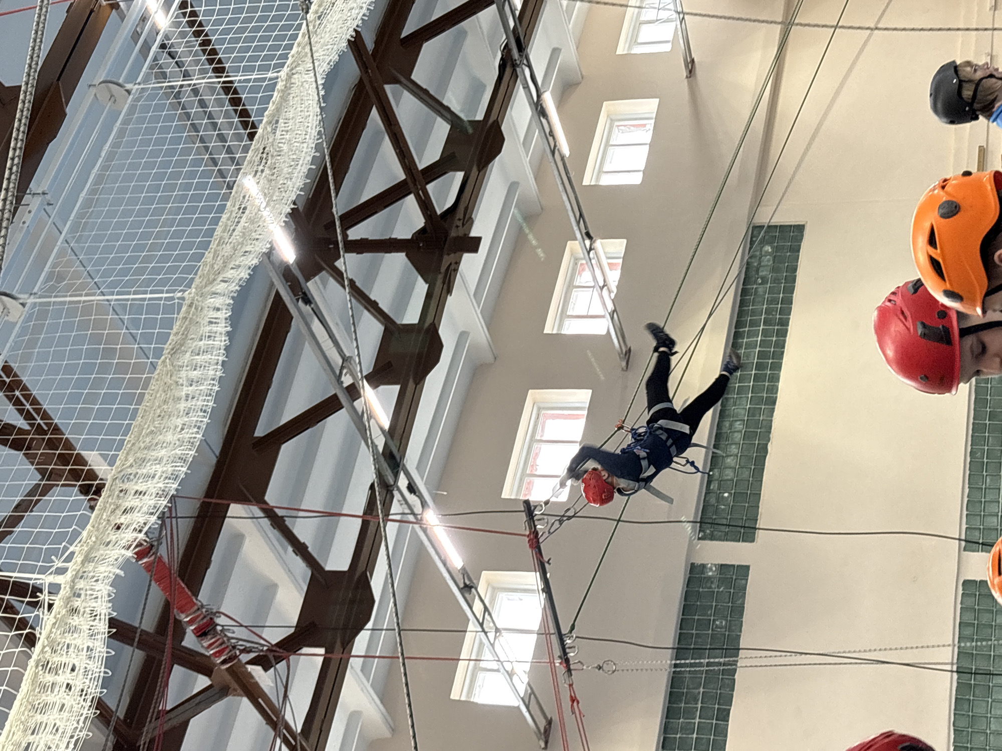 A view, tilted slightly to the left, shows a group of individuals participating in a high ropes course inside a building. The individuals are wearing helmets, some in red, others in orange and black. They are suspended by safety ropes and harnesses from the ceiling infrastructure. One person is positioned mid-air, dressed in dark-colored attire, connected to the ropes system, with their face obscured by a red helmet. The architectural backdrop includes a grid-like net on the left, and a wall with regularly spaced windows on the right. The overall color scheme is dominated by whites, grays, and muted tones, suggesting an indoor gymnasium or recreational center environment. The building is located in the city of Berezniki, Russia.
