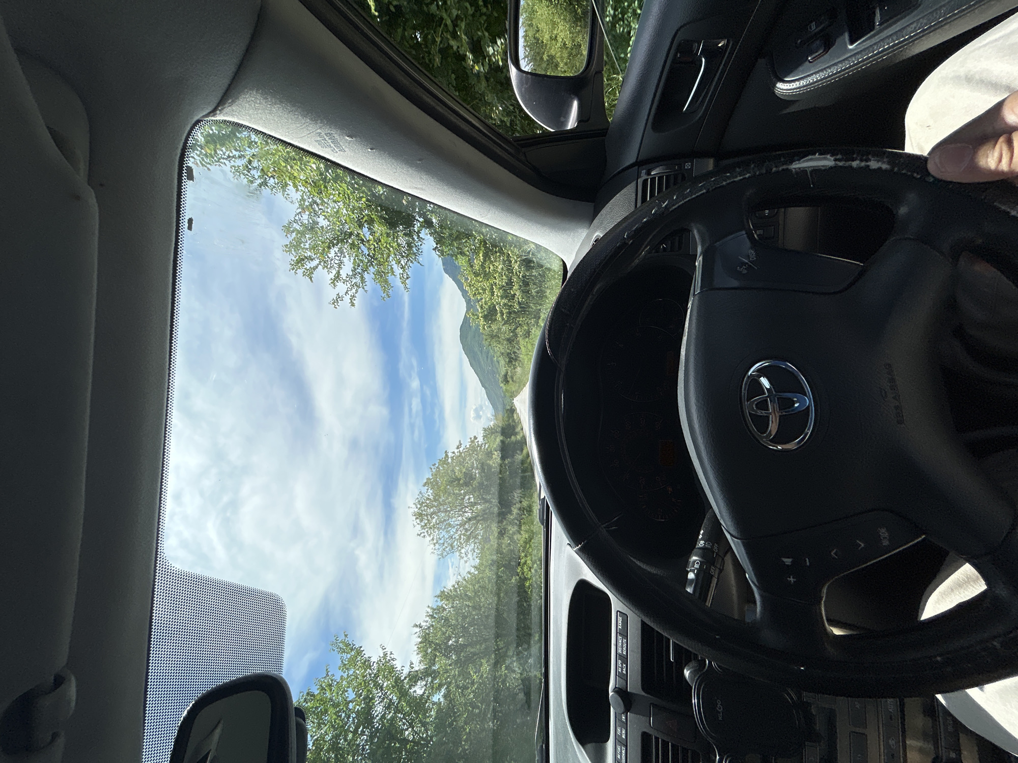 The image depicts a view from inside a Toyota vehicle, likely a minivan, driving on a road in a rural or mountainous area near Glozhene, Bulgaria. The driver's hand is visible on the worn steering wheel. The dashboard and center console are also visible. The view through the windshield shows a partly cloudy sky, trees lining the road, and a mountain in the distance. The side mirror is also visible, reflecting the surrounding greenery.