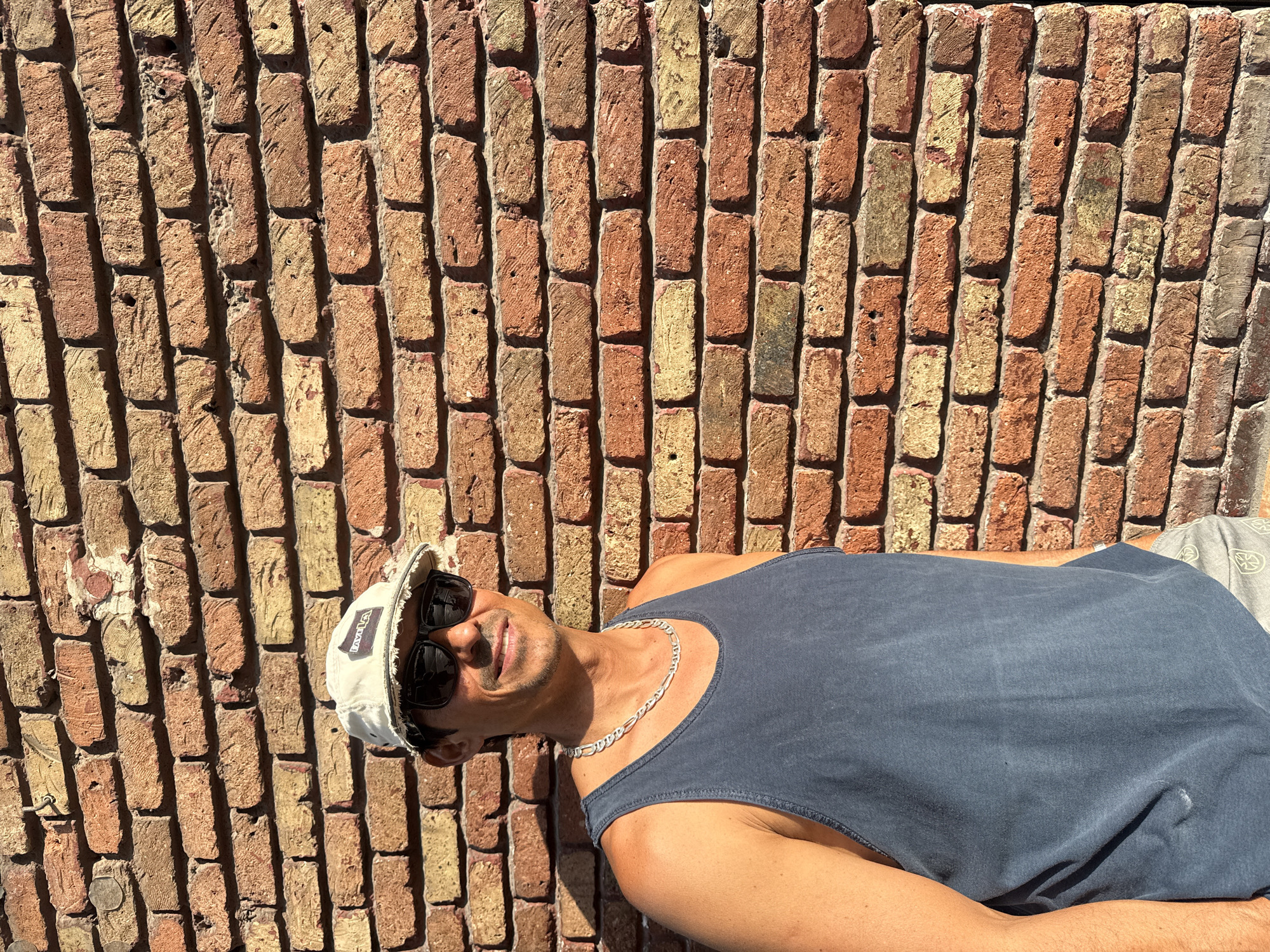The media file shows a man lying against a brick wall in Barcelona, Spain. The man is wearing a dark blue tank top, a white baseball cap, and sunglasses. He has a silver chain around his neck. The brick wall is made of weathered red and yellow bricks, with visible mortar. Sunlight is casting shadows on the wall, suggesting it is daytime, likely sunny. The man appears relaxed and is smiling. Some faint text is visible on the brim of his hat, appearing to read "LAVAL." The overall scene evokes a casual, warm-weather atmosphere.