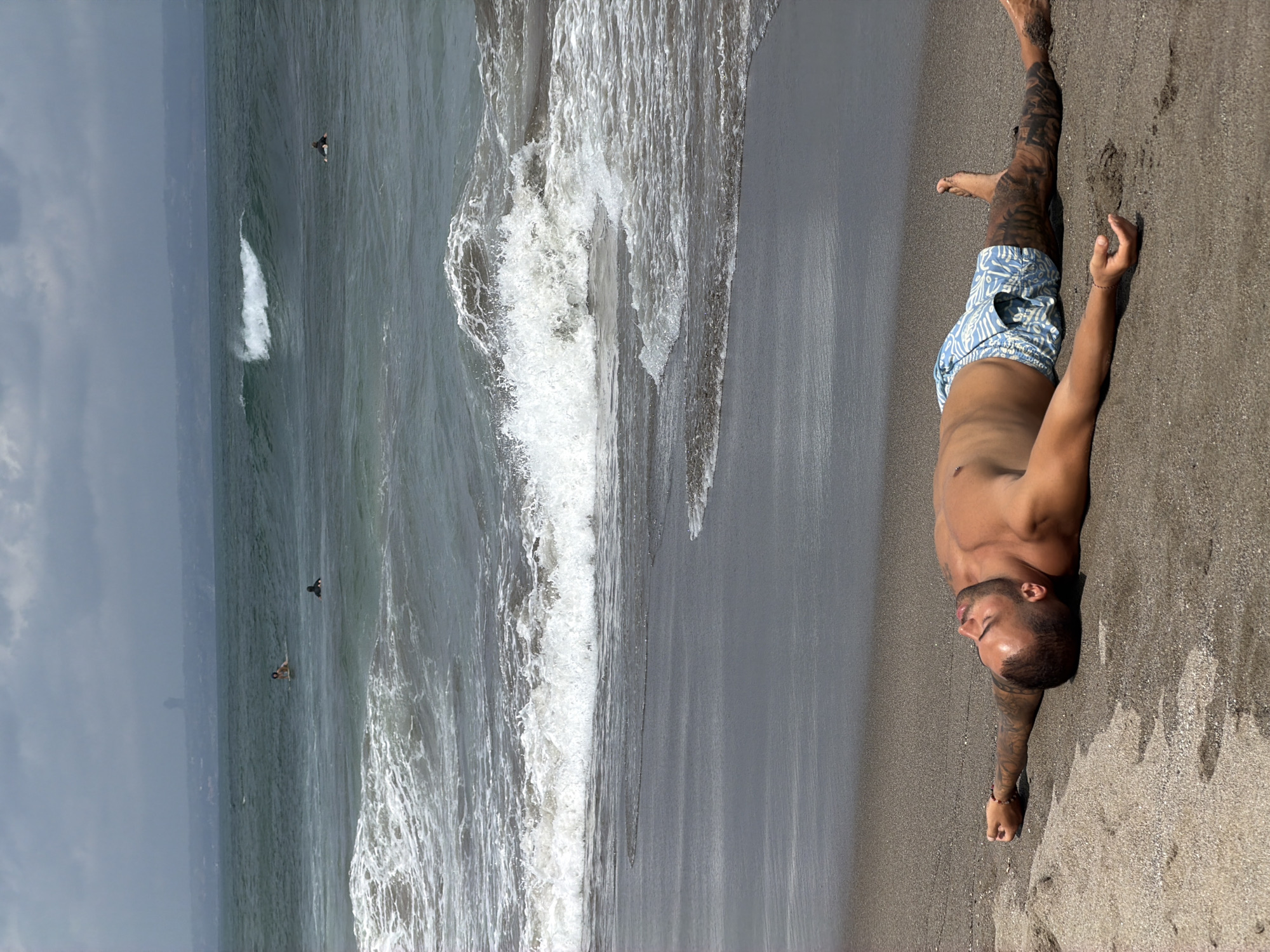 A man with dark hair, a beard, and extensive tattoos on his arms and legs is lying supine on a dark sand beach. He is wearing blue and white patterned swim trunks. His arms are outstretched to his sides, and his eyes are closed. The ocean, with moderate waves and a cloudy sky, is visible in the background. Several surfers are in the water further out. The scene appears to be in North Kuta, Indonesia.