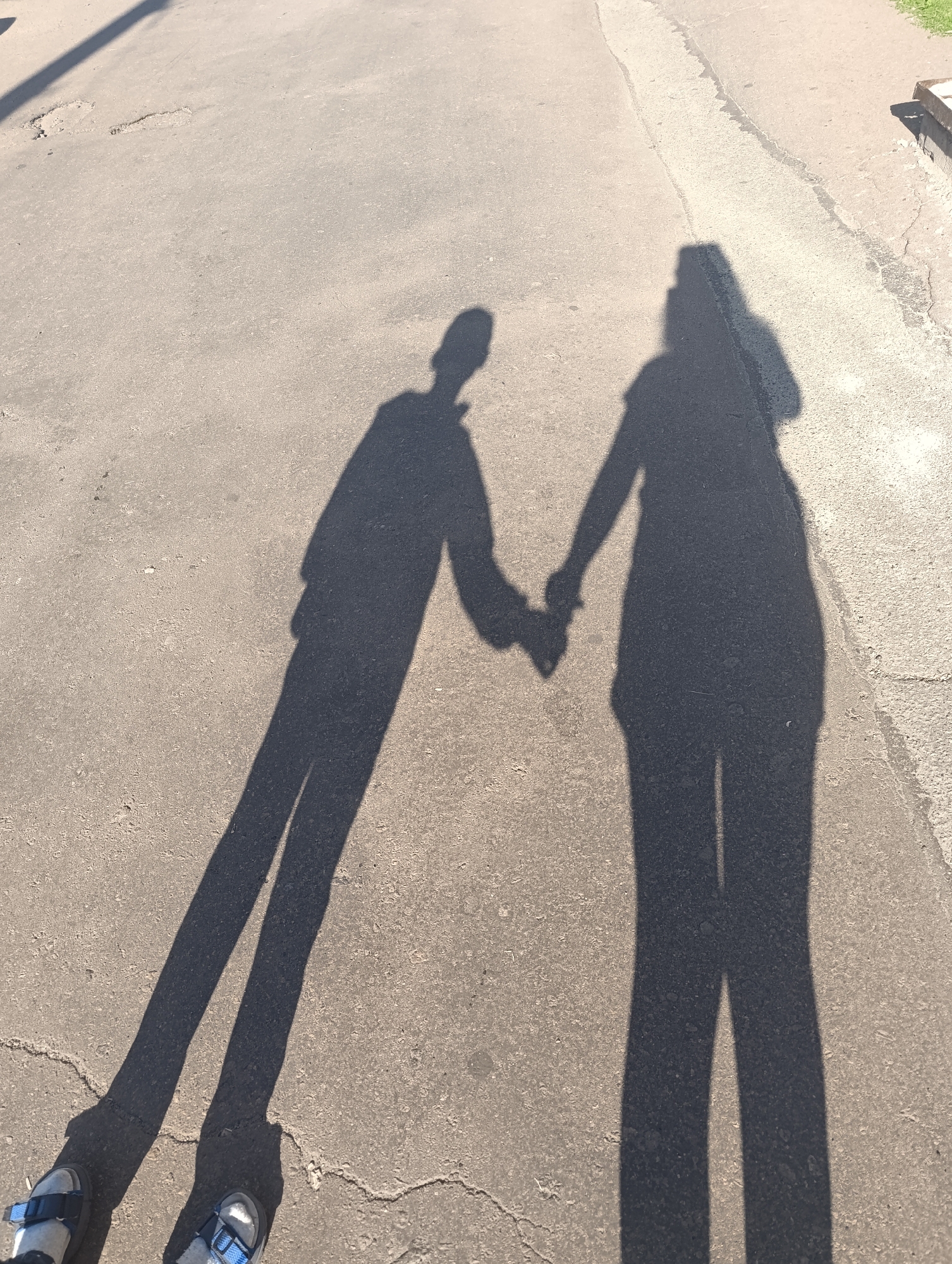 The image shows two shadows on an asphalt surface. One shadow is of a person wearing a long-sleeved shirt and pants. The other shadow is of a person wearing a dress or long shirt and pants. The two shadows are holding hands. The shadows are cast by two people standing on a paved area, possibly a street or sidewalk, in Chernihiv, Ukraine. The ground is asphalt with some cracks and debris. The lighting suggests it is daytime, with the sun casting long shadows. In the foreground, the shadows of two pairs of feet wearing sandals are visible.