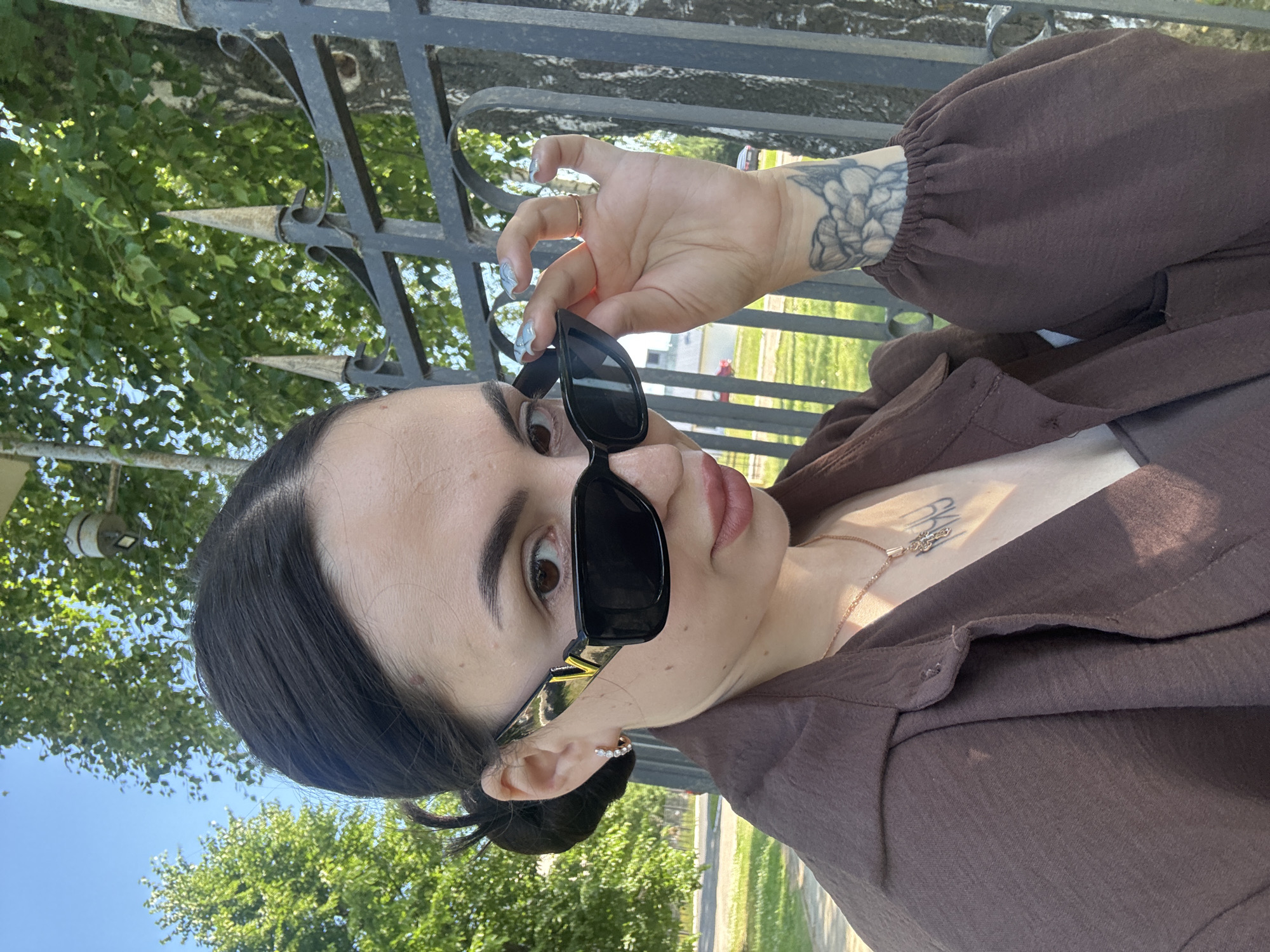 In Chaikovsky, Russia, a young woman with dark hair pulled back poses for a selfie outdoors. She is wearing oversized black sunglasses with a gold "V" emblem on the temples, a brown collared shirt with puffed sleeves, and a delicate gold necklace with a cross pendant. Her right hand is raised, adjusting her sunglasses, revealing manicured nails and a gold ring on her index finger. A tattoo of a floral design is visible on her left forearm. The background shows a black metal fence with decorative pointed finials, lush green trees, and a clear blue sky. The lighting suggests it is a sunny day.