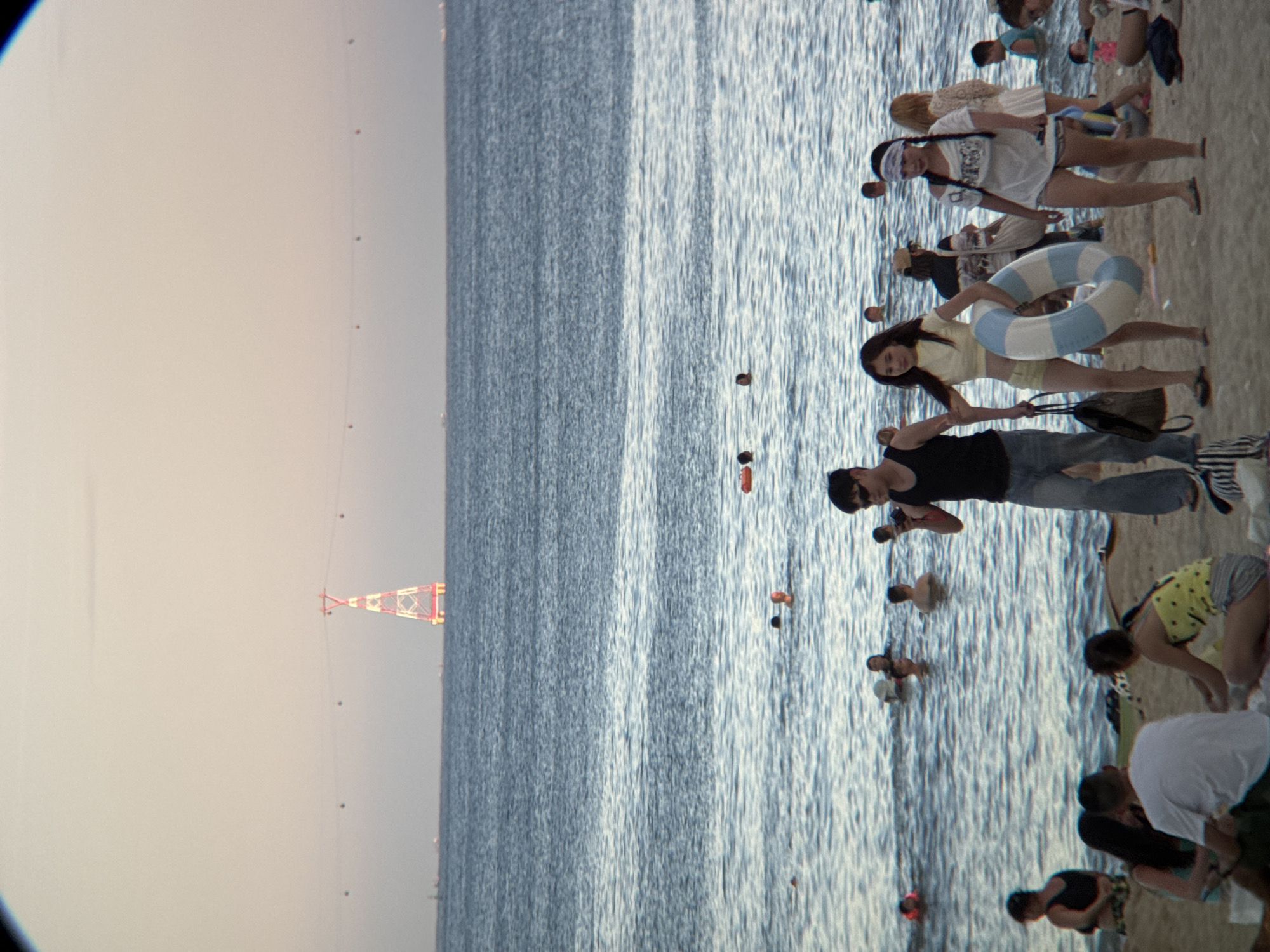 The image depicts a beach scene with numerous people in the water and on the sand, suggesting a public beach. In the background, a distinctive red and white pylon, likely part of a cable car system, is visible over the ocean. This pylon, identified as part of the Nha Trang cable car system connecting to Vinpearl Land, and the overall setting of a coastal city with a beach are characteristic of Nha Trang, Vietnam. The water appears calm, with many individuals swimming or wading. On the shore, several groups and individuals are present, some sitting, others standing. Among them, a man is photographing a woman carrying a blue and white striped life preserver, while another woman stands nearby. The atmosphere suggests a recreational day at the beach.