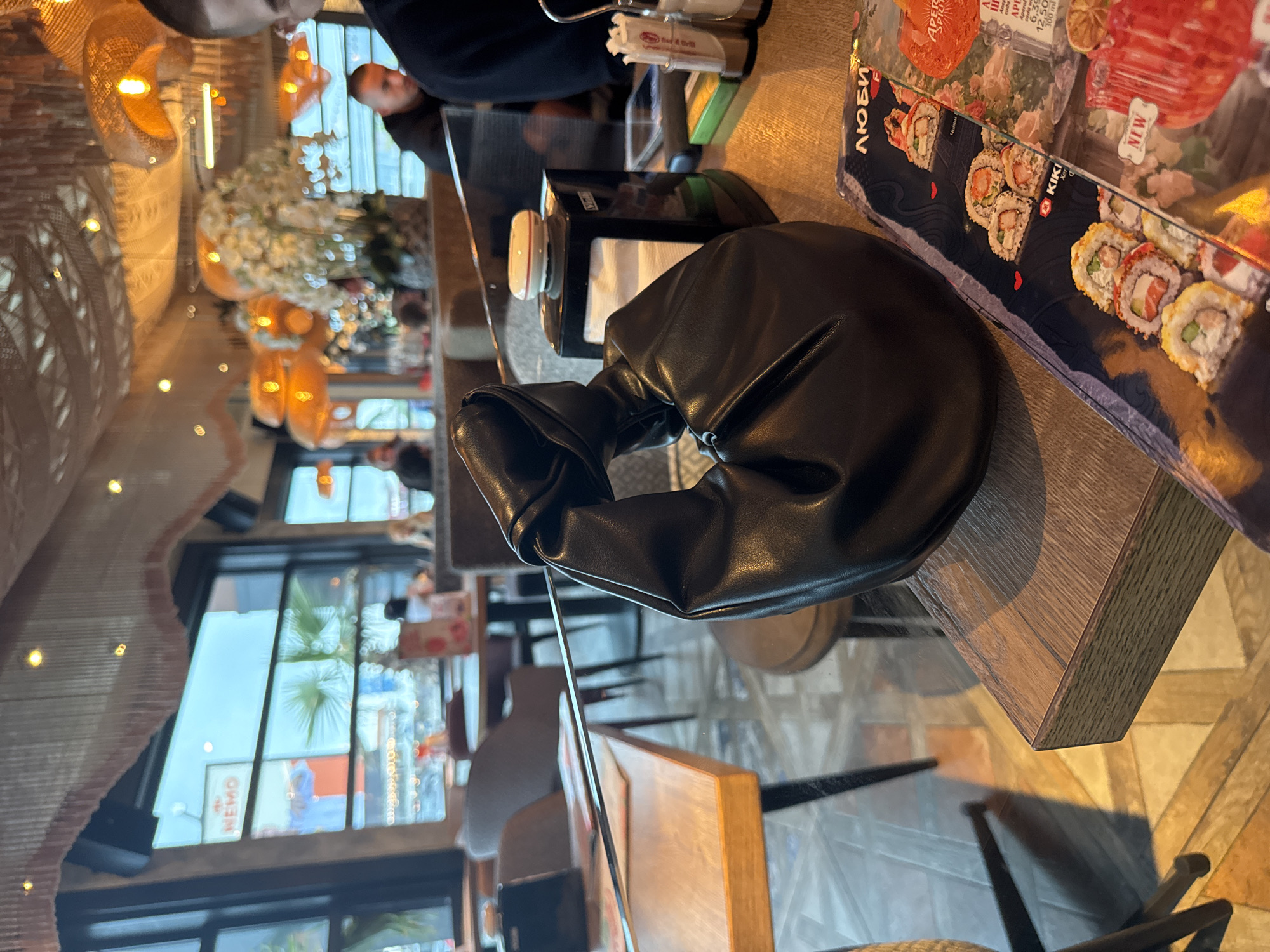 The image depicts a casual indoor dining scene, likely in a restaurant or cafe in Varna, Bulgaria. In the foreground, a stylish black faux leather purse with a knotted handle rests on a light-colored wooden table. To its right, a colorful sushi menu, possibly under a clear cover, displays various sushi rolls. Visible text on the menu includes "ЛЮБИ" (LUBI), "KIKI," "NEW," "APEROL SPRITZ," and "APPLE JUICE."
Behind the purse, a black napkin dispenser and a white salt shaker, potentially branded "Salt & Drill," are also on the table.
The background reveals a warm and inviting restaurant interior. Decorative light fixtures with woven shades cast a soft glow, complementing ambient ceiling lights. A large, elegant floral arrangement, appearing to be white roses, adds a touch of sophistication. Through expansive windows on the left, natural daylight illuminates the space, and reflections show a sign with the text "NEMO" and hints of outdoor greenery. Several patrons are visible in the blurred background, some seated at tables, suggesting a lively but relaxed atmosphere. One individual's face is partially visible in the upper right. The flooring features a geometric patterned tile design. The scene captures a moment of leisure, likely during daytime.