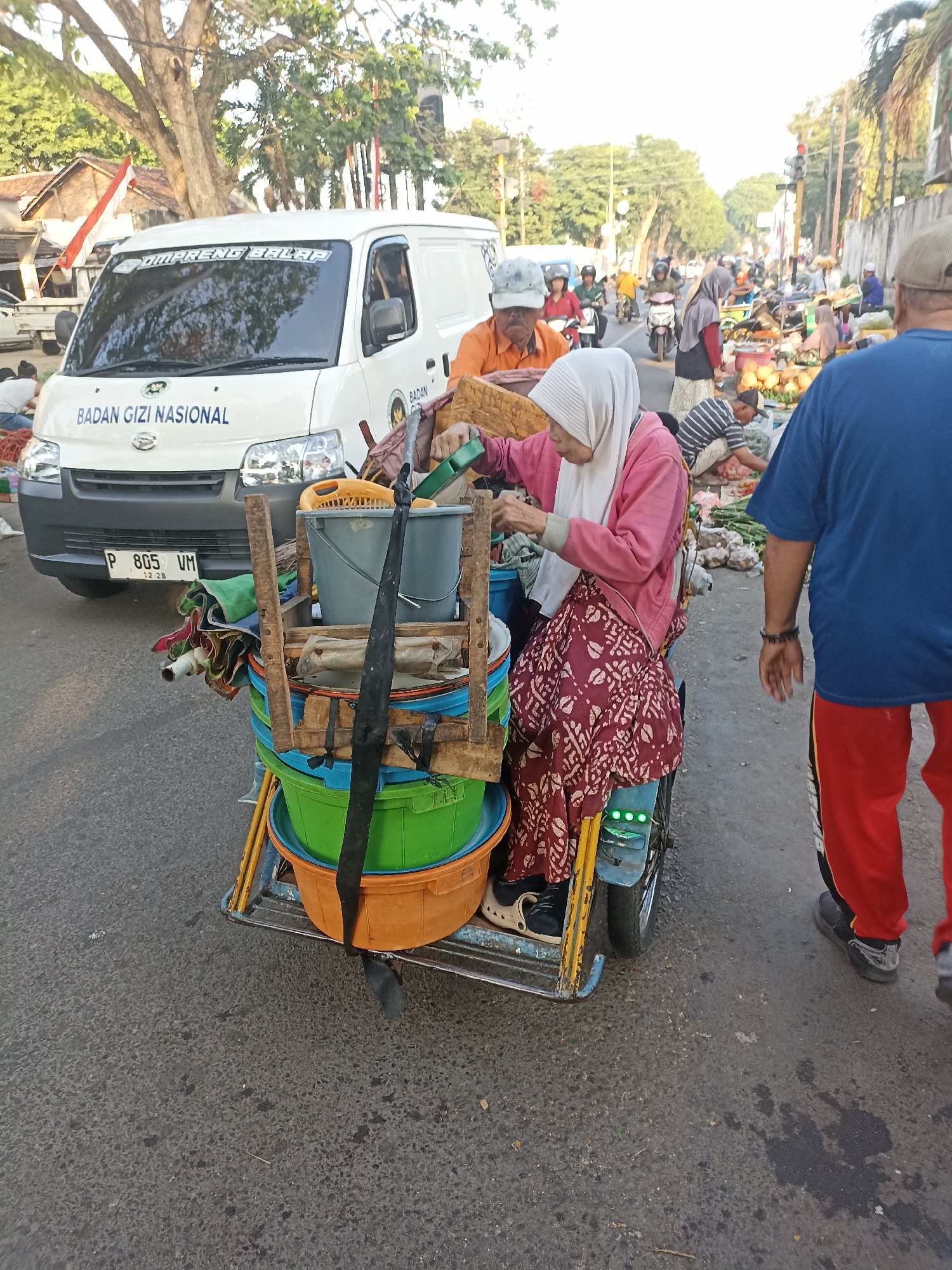 The image depicts a street scene, likely a market area, in Kelurahan Kepatihan, Indonesia. In the foreground, a woman wearing a pink hijab and a patterned maroon skirt is seated on a motorized tricycle laden with stacked buckets and containers. She is actively engaged in sorting or preparing items from one of the buckets. To her right, a person wearing a blue shirt and red pants walks away from the camera. Behind the tricycle, a white van with the text "BADAN GIZI NASIONAL" is parked. The background shows a busy street with more people, other vehicles including motorcycles, stalls displaying produce, and trees lining the road. The lighting suggests it is daytime, likely morning or late afternoon, with sunlight filtering through the trees. The overall atmosphere appears to be that of a bustling marketplace or street vendor environment.