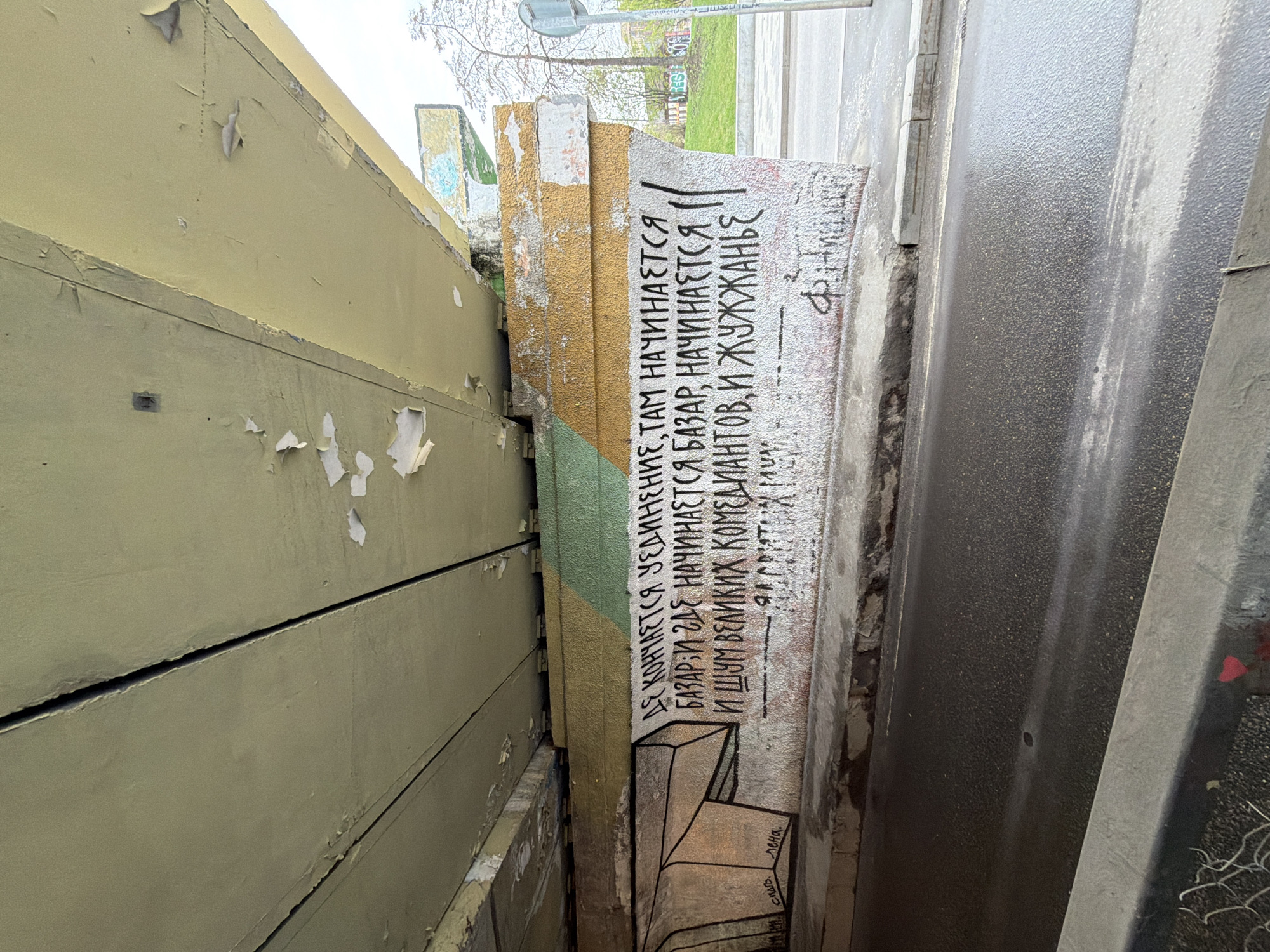 An oblique, downward-facing perspective captures a narrow urban passage in Moscow, Russia, between a building and a roadway. On the left, a light yellow or khaki-colored building wall shows signs of age with extensive peeling paint and visible horizontal seams.
In the center, a robust concrete or stone barrier separates the building from the road. This barrier is adorned with graffiti featuring black text on a textured white and gold background. The visible Russian text, clearly rendered, reads: "ГДЕ КОНЧАЕТСЯ УЕДИНЕНИЕ, ТАМ НАЧИНАЕТСЯ БАЗАР И ГДЕ НАЧИНАЕТСЯ БАЗАР, НАЧИНАЕТСЯ И ШУМ ВЕЛИКИХ КОМЕДИАНТОВ, И ЖУЖЖАНЬЕ" (Where solitude ends, there begins a bazaar, and where a bazaar begins, there begins the noise of great comedians, and buzzing). Below the main text, a small drawing depicts geometric shapes, possibly abstract steps or boxes, and an apparent signature or attribution "Ф. Достоевский" (F. Dostoevsky) referring to the quote's author.
To the right, a dark grey asphalt surface, possibly a road or sidewalk, appears wet and reflects the dim, overcast sky seen in the background. A blurred view beyond the road reveals bare tree branches, suggesting late autumn, winter, or early spring, and a glimpse of a street sign pole. No people or specific activities are depicted, focusing instead on the architectural and textual elements of the urban environment. The scene conveys a sense of everyday city grit and intellectual graffiti.