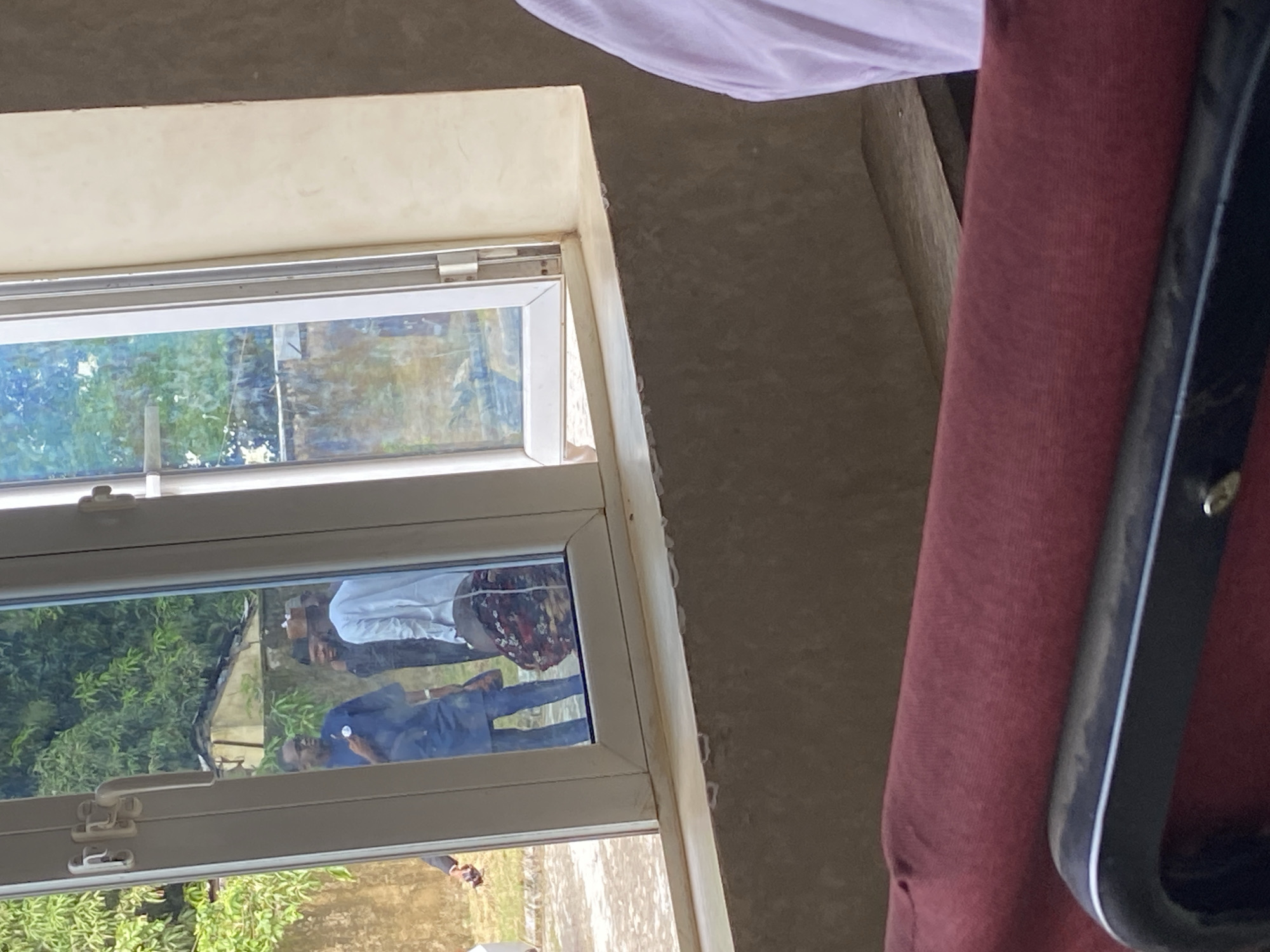 The image depicts an interior view looking out through a window onto an outdoor scene in Mararaba, Nigeria. In the immediate foreground, the top of a dark red upholstered chair or seat is visible, with a black and silver armrest spanning the lower part of the frame. To the right, a portion of a light purple garment or fabric is visible, suggesting a person is seated or standing just outside the frame to the right.
The central feature is a white-framed casement window, partially open on the right pane, offering a clear view of the exterior. Below the window is a simple windowsill, appearing to be concrete or plaster, with some minor surface irregularities. The interior walls surrounding the window are a light, muted tone, possibly unpainted or minimally finished.
Outside the window, a group of individuals is visible on what appears to be a paved or graveled ground surface. Three men are prominent:
1. On the left, a man wearing a dark blue tunic and trousers, with glasses, holds a dark object (possibly a mobile phone or communications device) to his right ear. A light-colored badge or emblem is visible on his left chest area. He faces slightly towards the right.
2. To his right, partially obscured, is a man in dark clothing, looking towards the right.
3. Further right, a man in a light blue or white tunic and a light brown cap (possibly a Kufi or similar headwear) stands, facing slightly towards the left.
4. Below the man in the light tunic, the lower body of a person is visible, wearing a dark skirt or wrapper with a floral or intricate pattern in red, white, and other colors.
5. A partial view of an arm and hand holding a dark object is visible on the far left edge of the window, suggesting another person is present.
The background outside the window features lush green foliage, indicative of trees or dense bushes. A section of a light-colored wall or fence is visible, with what appears to be the roofline or structure of a small, rustic building behind it, made of earth-toned materials. The overall scene suggests a casual gathering or interaction occurring outdoors on a clear day.
