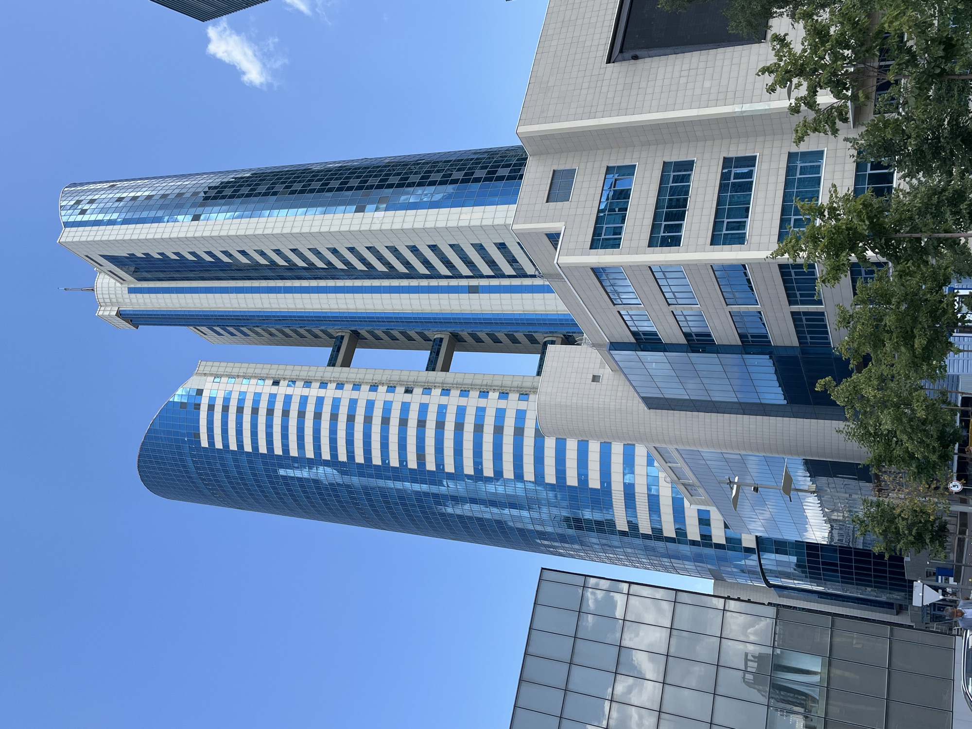 In Astana, Kazakhstan, a cluster of modern skyscrapers is captured under a clear blue sky. The tallest buildings are characterized by their glass facades, featuring alternating blue and white panels that reflect the sky. One tower has a curved, organic shape, while another is more rectilinear, connected by an elevated walkway. In the foreground, a lower, angular building with reflective glass windows displays clouds. Mature green trees line the base of the structures, and a glimpse of a sidewalk and street is visible. A few people are present on the sidewalk, one of whom is walking. The scene conveys a sense of urban development and architectural ambition.