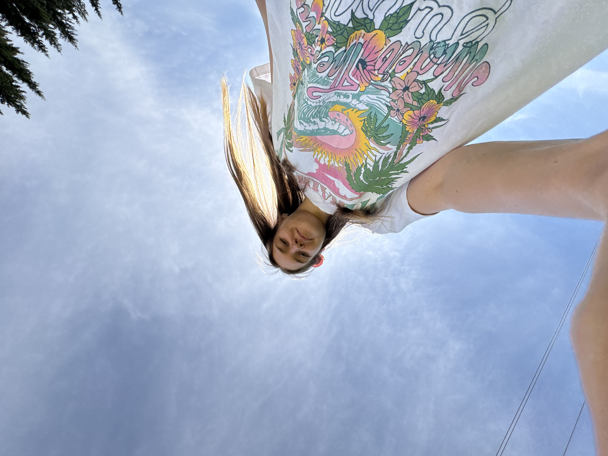 A young woman is pictured from a low angle, looking down at the camera. She has long brown hair with golden highlights. She's wearing a white t-shirt with a colorful graphic print. The background is a cloudy sky, with the top of a tree visible in the upper left corner. Power lines are also visible in the lower right corner. The location is presumed to be outside. The city is indicated as Sibir', Russia but there are no visual elements to confirm that.