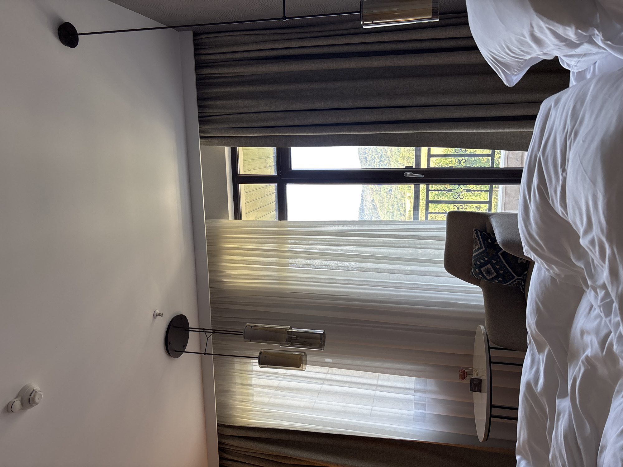 The image shows an interior view of a hotel room, likely in Razliv, Bulgaria, looking towards a balcony and a landscape. In the foreground, a rumpled white duvet and pillow suggest a recently used bed. To the left, a small round table with a vase holding a single red flower and a QR code is visible. Behind it, a comfortable armchair with a patterned cushion is placed.
The room features large windows covered by sheer white curtains and heavier dark grey drapes. Through the window, a balcony with a metal railing is visible, beyond which is a view of rolling green hills and mountains under a bright sky. Two modern pendant lights hang from the ceiling. A smoke detector is mounted on the ceiling. The overall impression is of a well-appointed hotel room with a scenic view.
