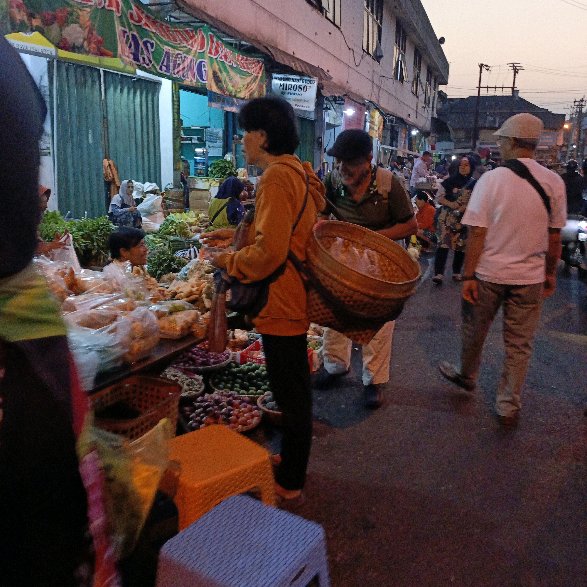 The image depicts a bustling street market scene in Salatiga, Indonesia. In the foreground, a person wearing an orange hoodie is seen from the waist down, carrying a large woven basket on their back. Another person in a white t-shirt and light-colored pants walks away from the camera, with a dark bag slung across their chest. The background reveals a row of market stalls with various goods displayed, including fresh produce such as onions and limes. People are seen browsing and interacting, indicating active commerce. Buildings line the street, and overhead power lines are visible against the twilight sky. The overall impression is of a lively and typical Indonesian market at dusk.