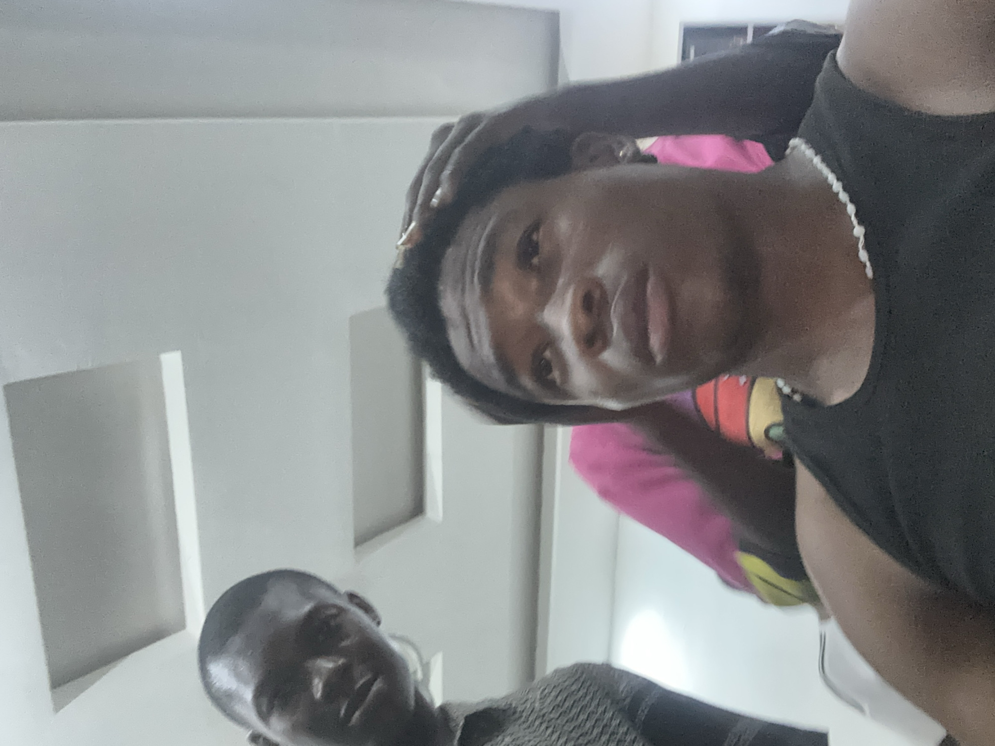 Two Black men are visible indoors. One man is facing the camera, wearing a black tank top and a beaded necklace, with a hand on his head. Another person, partially visible in the bottom left corner, is wearing a patterned shirt. The location is presumed to be Ogwashi-Uku, Nigeria. The environment appears to be an interior space with white walls.