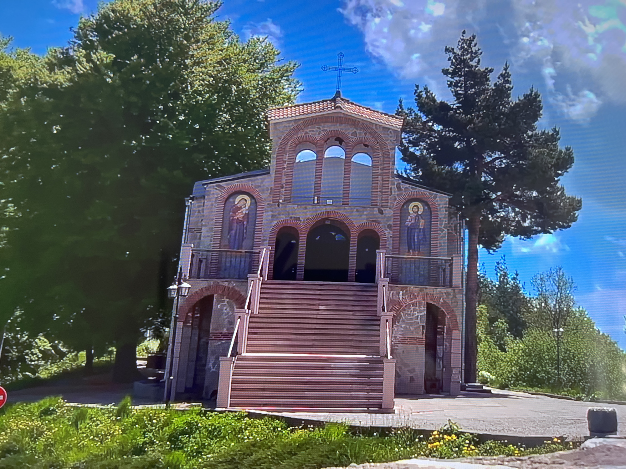 The image depicts a two-story church with a red-tiled roof and a prominent cross on top. The facade is constructed of stone and brick, featuring arched windows and doorways. Two religious icons are positioned on either side of the upper level. A wide set of stairs leads up to the entrance. The church is surrounded by lush greenery, including mature trees and flowering plants. The sky is blue with scattered clouds. The church is located in Unknown, Unknown.