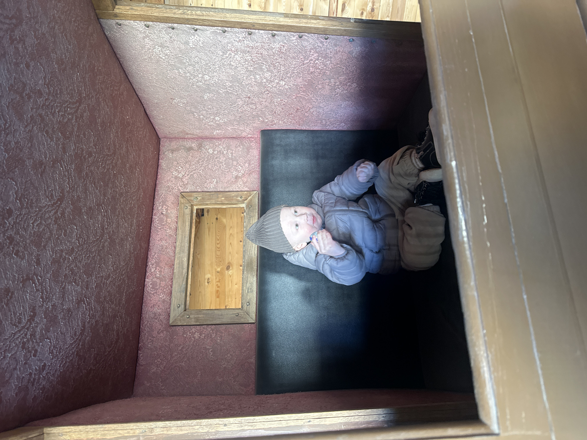 A small child in a grey winter outfit lies on a black cushioned bench inside a small room with pink patterned wallpaper. A small wooden window is on the left wall. The room appears to be a staged exhibit. No information in the image identifies Votkinsk, Russia.