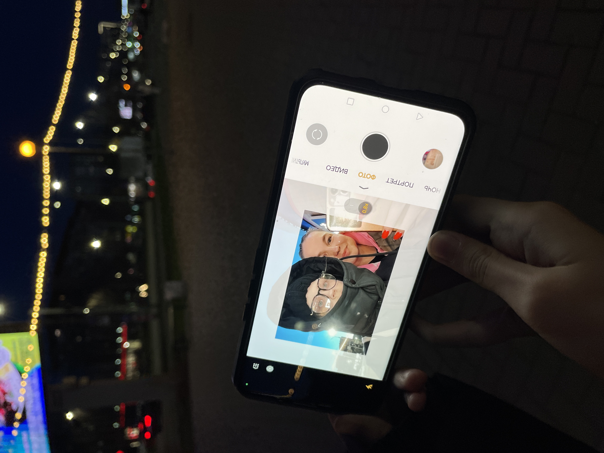 A person's hand holds a black smartphone in an indoor setting, looking out a window at night. The smartphone screen displays its camera application, with "ФОТО" (PHOTO) highlighted, indicating the current mode. Other visible camera modes include "ВИДЕО" (VIDEO), "ПОРТРЕТ" (PORTRAIT), "НОЧЬ" (NIGHT), and "МУЛЬТИ" (MULTI). Zoom options "0.8x" and "1x" are also visible.
On the phone's screen, a live preview or a recent photo shows two individuals. One person, closer to the viewer, wears a black beanie and glasses, looking directly at the camera with a slight smile. Behind them, another person with blonde hair and a pink top smiles broadly. The background within the phone's camera view is brightly lit, likely an indoor space.
Outside the window, in the background of the main image, is a blurred cityscape at night. Distant city lights create a bokeh effect, and a prominent vertical string of warm yellow lights, possibly a decorative street light or building facade, runs along the left side of the frame. The overall scene suggests someone is either taking a selfie or a portrait photo with the urban night lights as a backdrop, in Izhevsk, Russia. The image has a dark, nocturnal ambiance, primarily lit by the smartphone screen and the distant city lights.