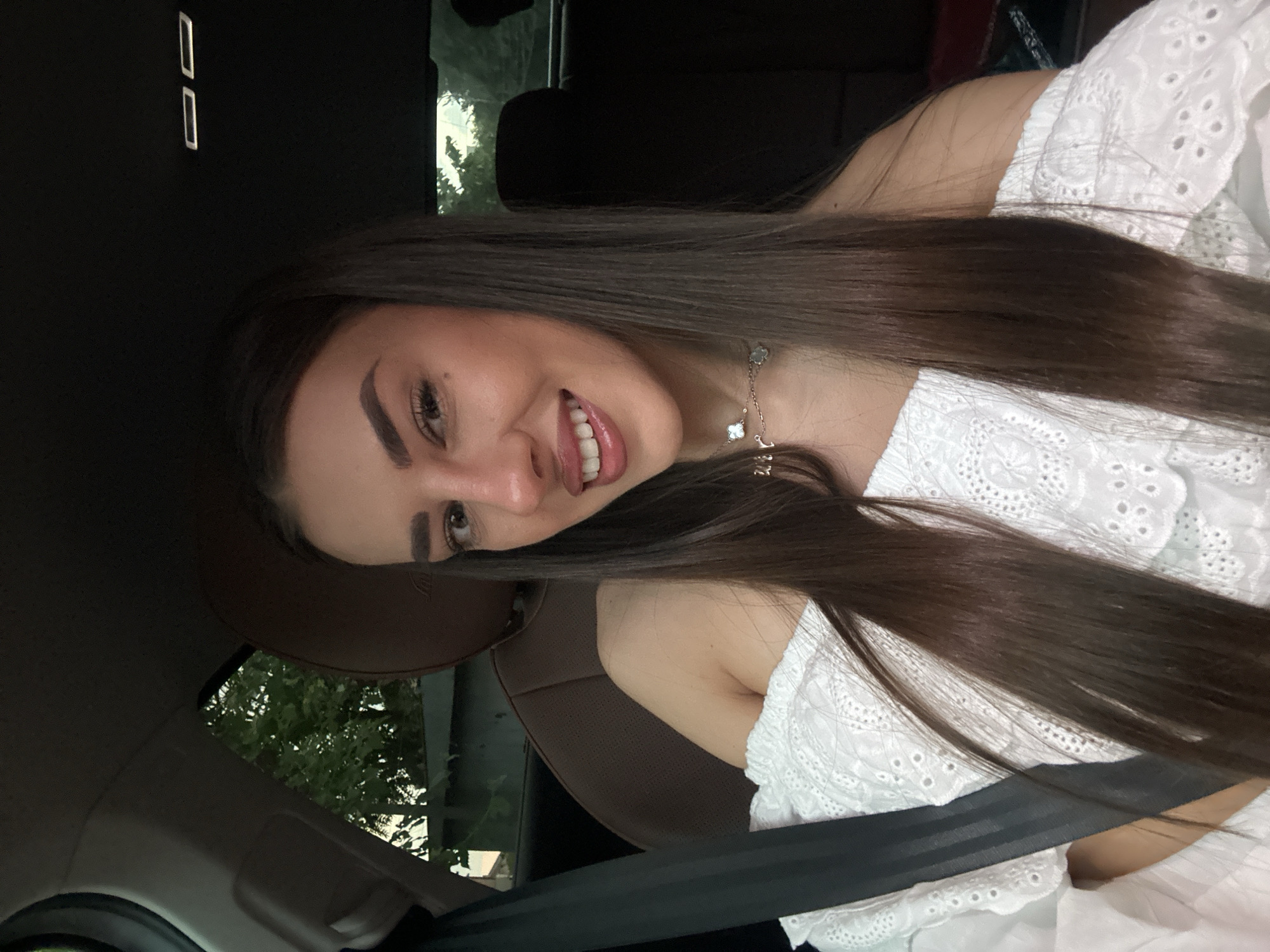 The image shows a young woman with long, dark brown hair, sitting in the passenger seat of a car. She is wearing an off-the-shoulder white top with intricate lace detailing and a delicate silver necklace. She is smiling, with her mouth slightly open, revealing her teeth. The interior of the car is visible, including the dark upholstery of the seats and a seatbelt across her chest. The background outside the car window shows trees and greenery, suggesting an outdoor setting, possibly on the outskirts of Petrich, Bulgaria, given the context. The lighting is natural, indicating it is daytime.