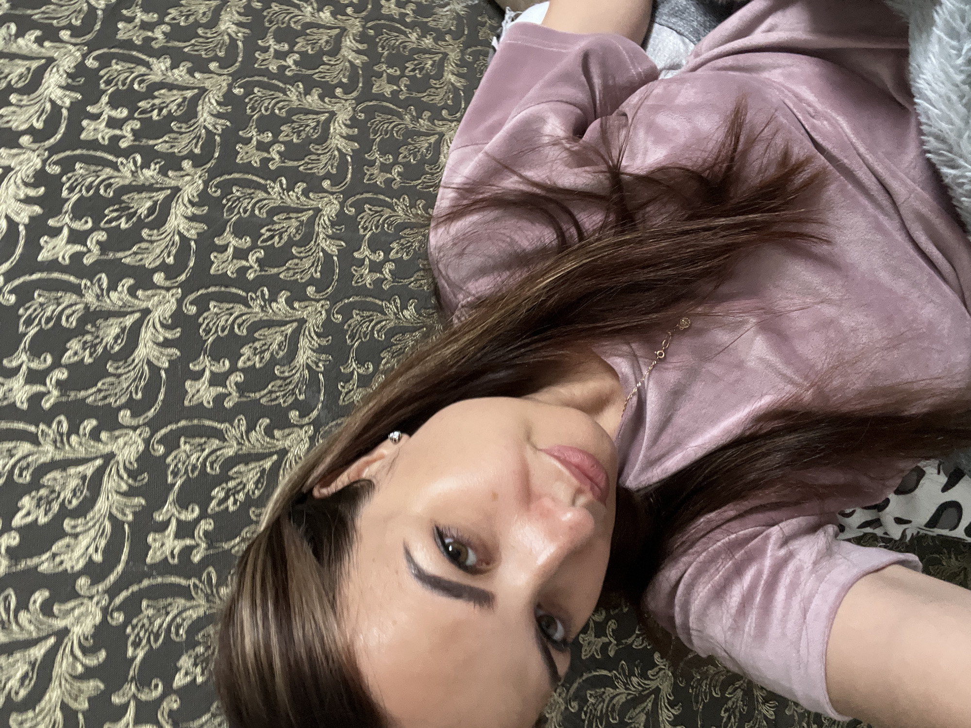 A medium shot, taken from a high angle, shows a woman lying on a patterned sofa. The woman has long, brown hair and is wearing a pink, velvety shirt. She is looking at the camera with her head tilted. She is wearing a small earring in her left ear. The sofa has a dark grey background with a gold, floral pattern. The woman is positioned on the left side of the frame.