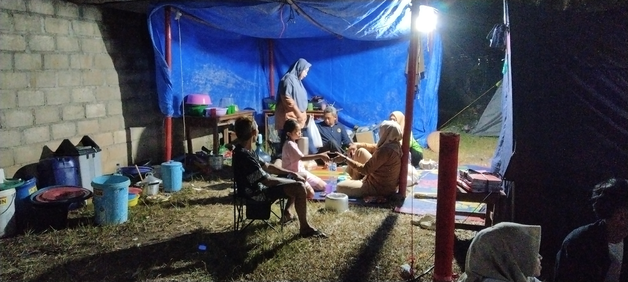 In Sido Rahayu, Indonesia, a group of people are gathered under a blue tarp shelter at night. The scene appears to be an outdoor gathering or temporary living space, possibly in a residential backyard or an informal settlement. The ground is covered in dry grass and dirt. Several large blue and white plastic buckets, as well as other containers and household items, are visible near a brick wall to the left of the shelter.
Inside the shelter, illuminated by a bright overhead light, several individuals are seated or standing. In the foreground, a man sits in a folding chair, facing away from the camera. To his right, a young girl in a pink shirt and pants sits on the ground. In the background, a woman wearing a grey hijab and a beige top stands, holding a white bag. Another man in a dark shirt is seated, and two women, one in a beige hijab and the other in a green shirt, are also seated. They are gathered around a low table or mat, suggesting a communal meal or activity. A tent structure is visible further back in the darkness.