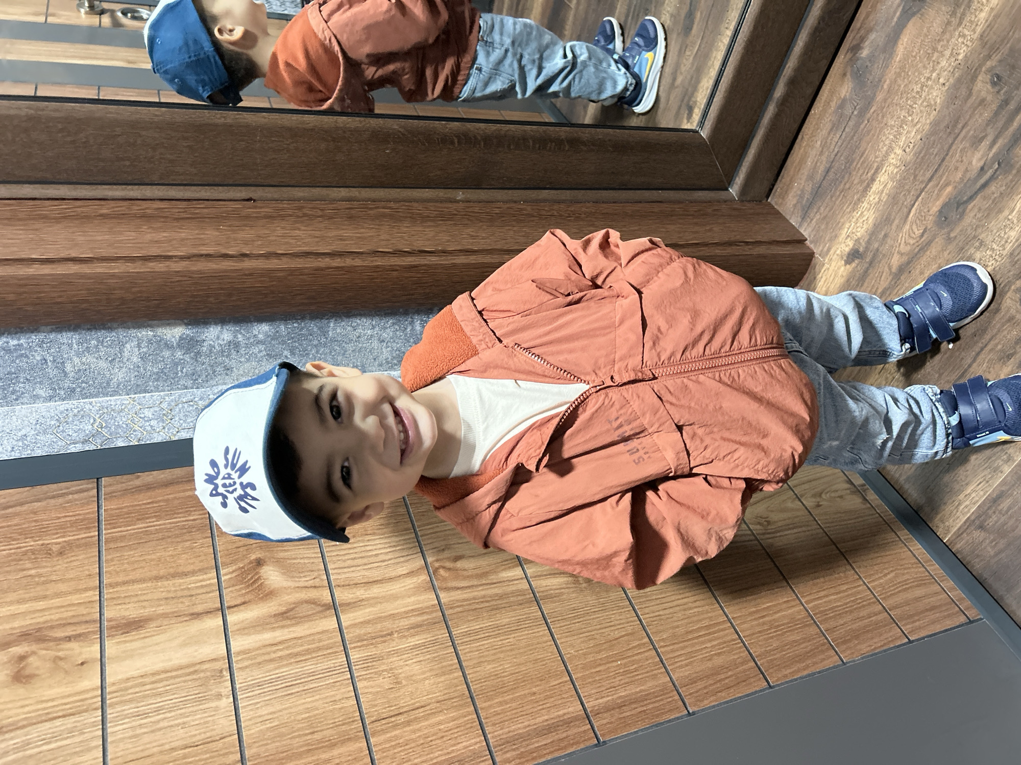 A young boy stands in front of a wooden door and a mirror. He is wearing a white t-shirt, an orange jacket with a zipper, light blue jeans, a white and blue baseball cap, and blue sneakers. The boy is smiling and looking at the camera. In the mirror, his reflection is visible, showing his back and the side of his face. The background consists of a wooden wall with horizontal planks and a grey textured wall. The floor is made of wooden planks. The scene appears to be indoors, possibly in a home or a fitting room. There is no information available to connect this image to the city of Kardzhali, Bulgaria.