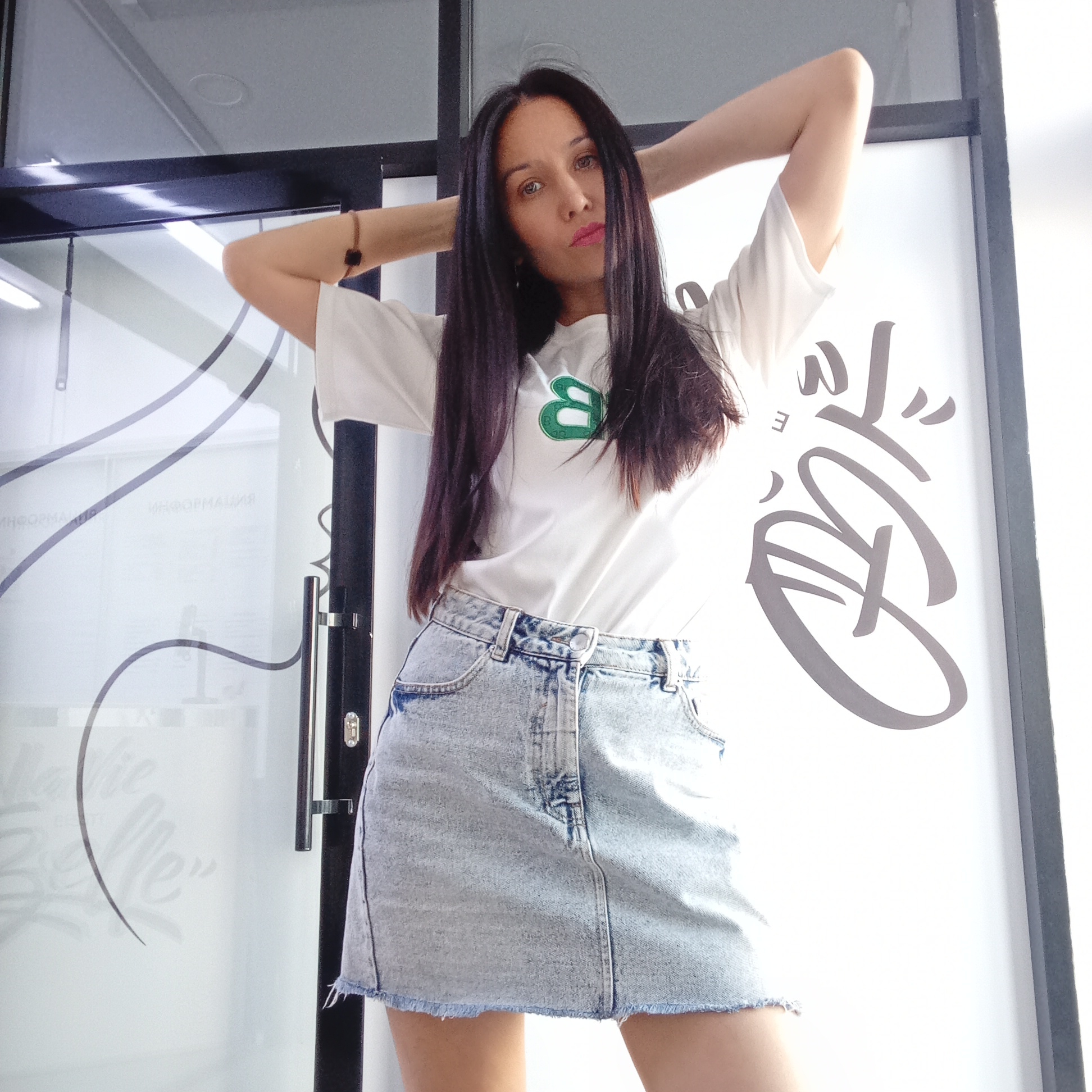 A woman stands indoors in what appears to be a commercial or office space. She has long, dark hair and is wearing a white t-shirt with a green letter "B" and a denim skirt. Her arms are raised behind her head. The background includes a glass door with design elements and lettering, along with visible door hardware. The location is Izhevsk, Russia based on lettering on the door in Russian.